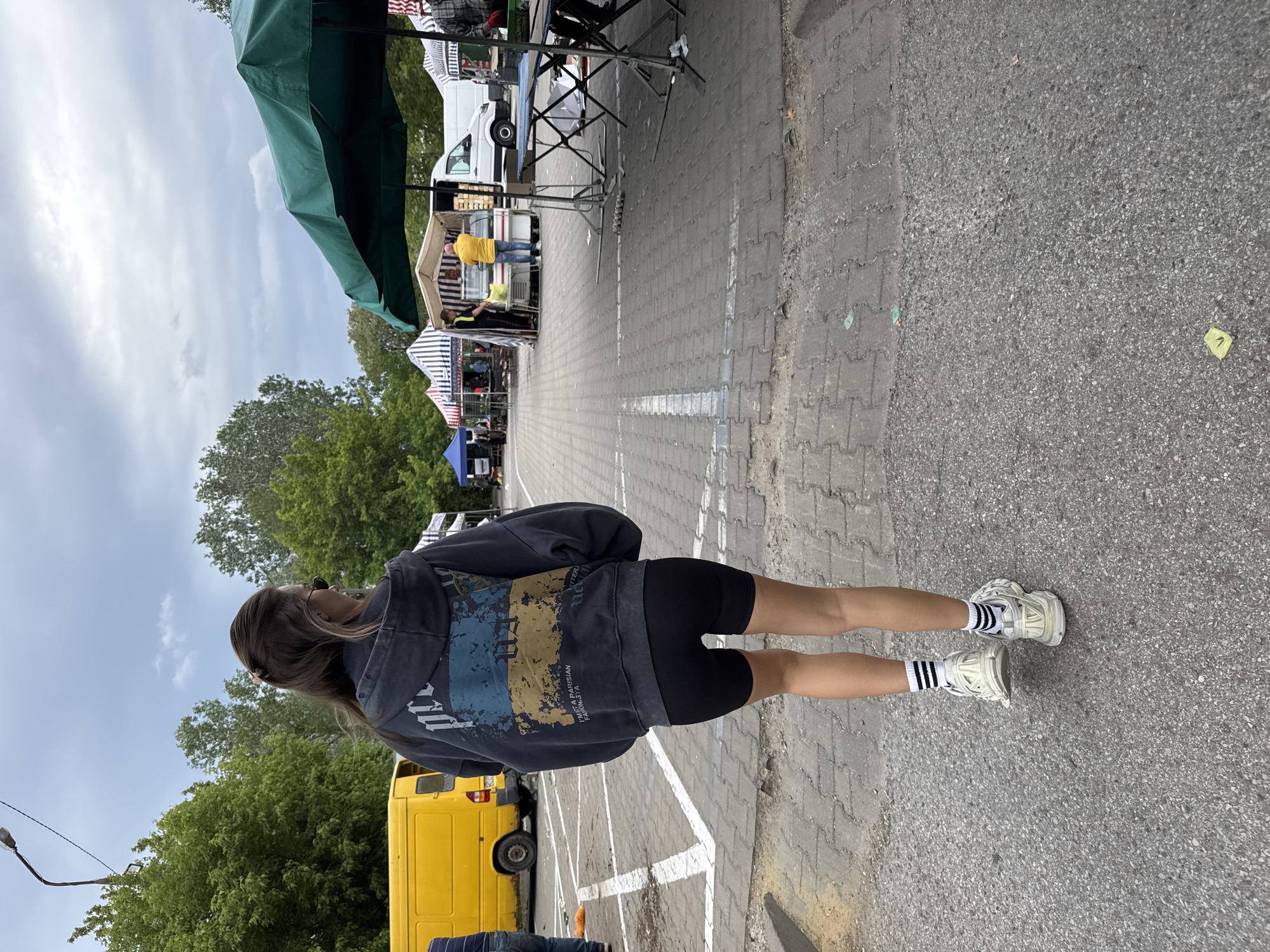 The image shows a woman with long hair, wearing a dark hoodie with the Ukrainian flag printed on the back, black cycling shorts, white sneakers, and black and white striped socks. She is standing on a paved area with white painted parking lines, facing away from the camera and toward a market or fair.
In the background, there are several vendor stalls with green and blue canopies. A white van and a yellow van are parked nearby. Trees line the background. The sky is cloudy. The location is an open space likely used for markets or events.