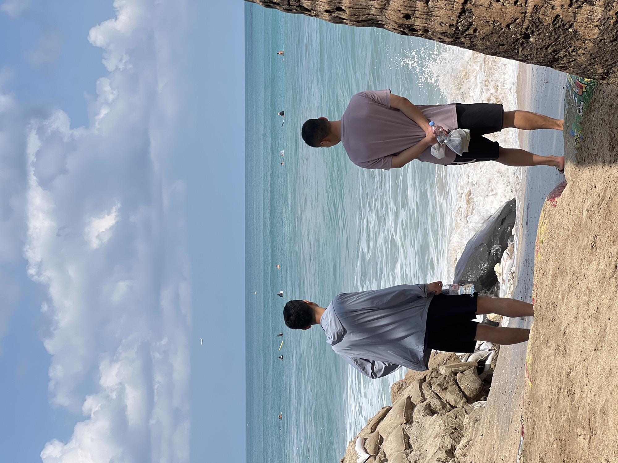 Two men stand on a sandy beach in Kuta, Indonesia, facing the turquoise ocean under a partly cloudy sky. The man on the left wears a grey hooded jacket and black shorts, holding a plastic bottle of water. The man on the right, with his hands clasped behind his back, wears a light purple t-shirt and black shorts, also holding a plastic bag containing items and a bottle. Several surfers are visible in the water, some on boards and others paddling. The shoreline is littered with sandbags and debris. An airplane is visible in the sky. A portion of a palm tree trunk is on the right edge of the frame.