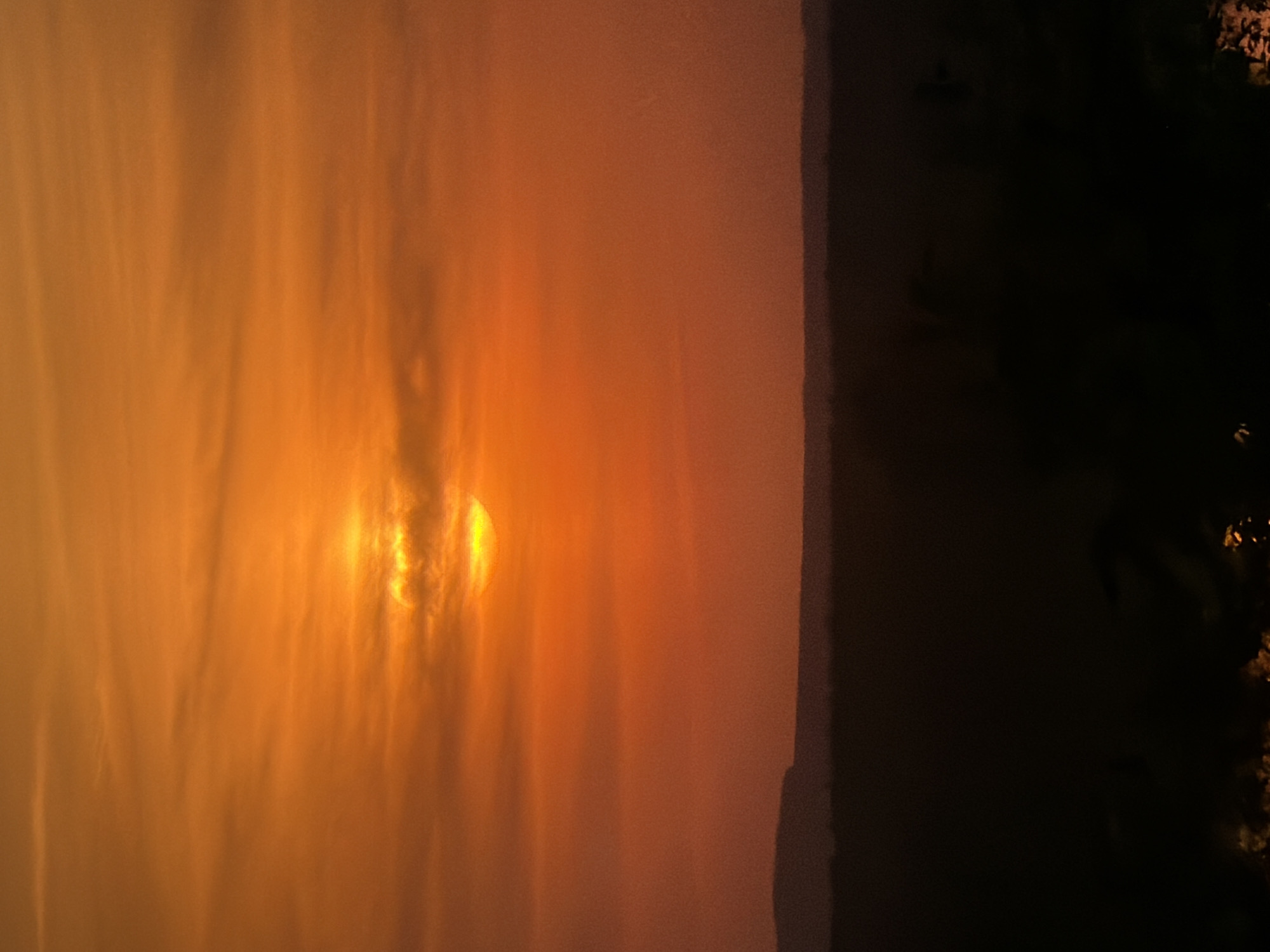 The image depicts a sunset over a hazy sky in Chaikovsky, Russia. The sun is visible as a bright, diffused orb through the haze, with orange and yellow hues dominating the sky. To the right, there's a dark area, possibly indicating trees or buildings, obscuring the horizon.