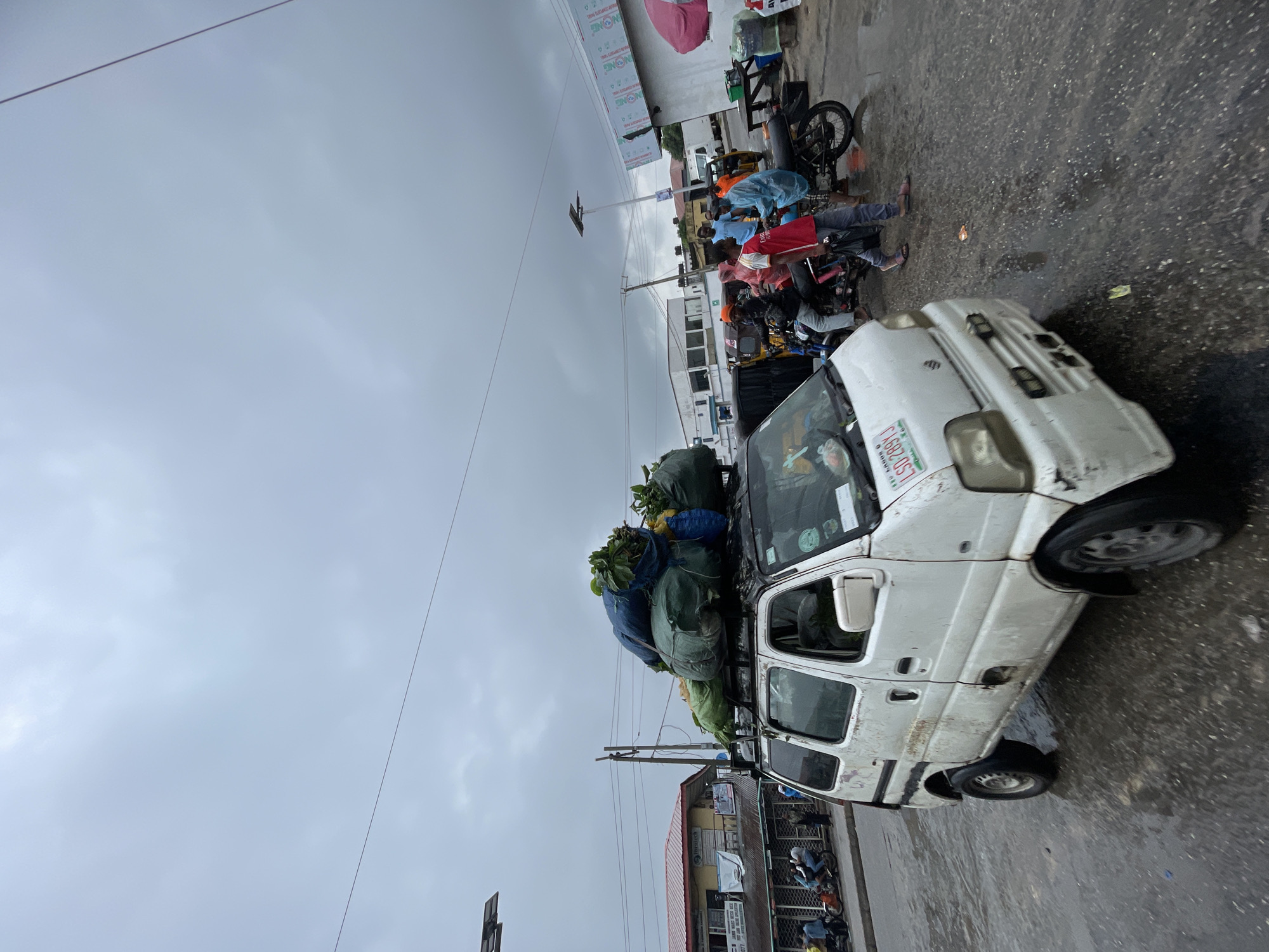 The image shows a white compact van overloaded with large bags of produce or goods strapped to its roof rack. The van is parked on a wet, paved surface, likely a street or roadside in Ado, Nigeria. The sky is overcast and gray, suggesting recent or ongoing rain. Behind the van, a row of motorcycles are parked, some with passengers wearing rain gear. Buildings and storefronts are visible in the background, indicating an urban or commercial area. There are also people gathered around the motorcycles. Powerlines stretch across the scene above.