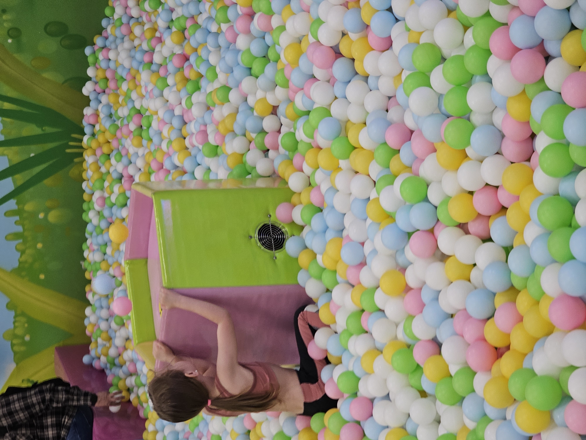 A high-angle indoor shot depicts a ball pit in Khimki, Russia. The pit is filled with colorful plastic balls: white, green, yellow, pink, and light blue. A multi-level play structure with green and pink panels rises from the ball pit. A young girl with brown hair tied in a ponytail is climbing into the structure. She wears a pink sports bra and black pants. At the edge of the frame, the bottom left, a person in a plaid shirt and jeans stands near the pit. The background features a mural with green trees against a blue sky. A ventilation fan is visible on the green structure panel.