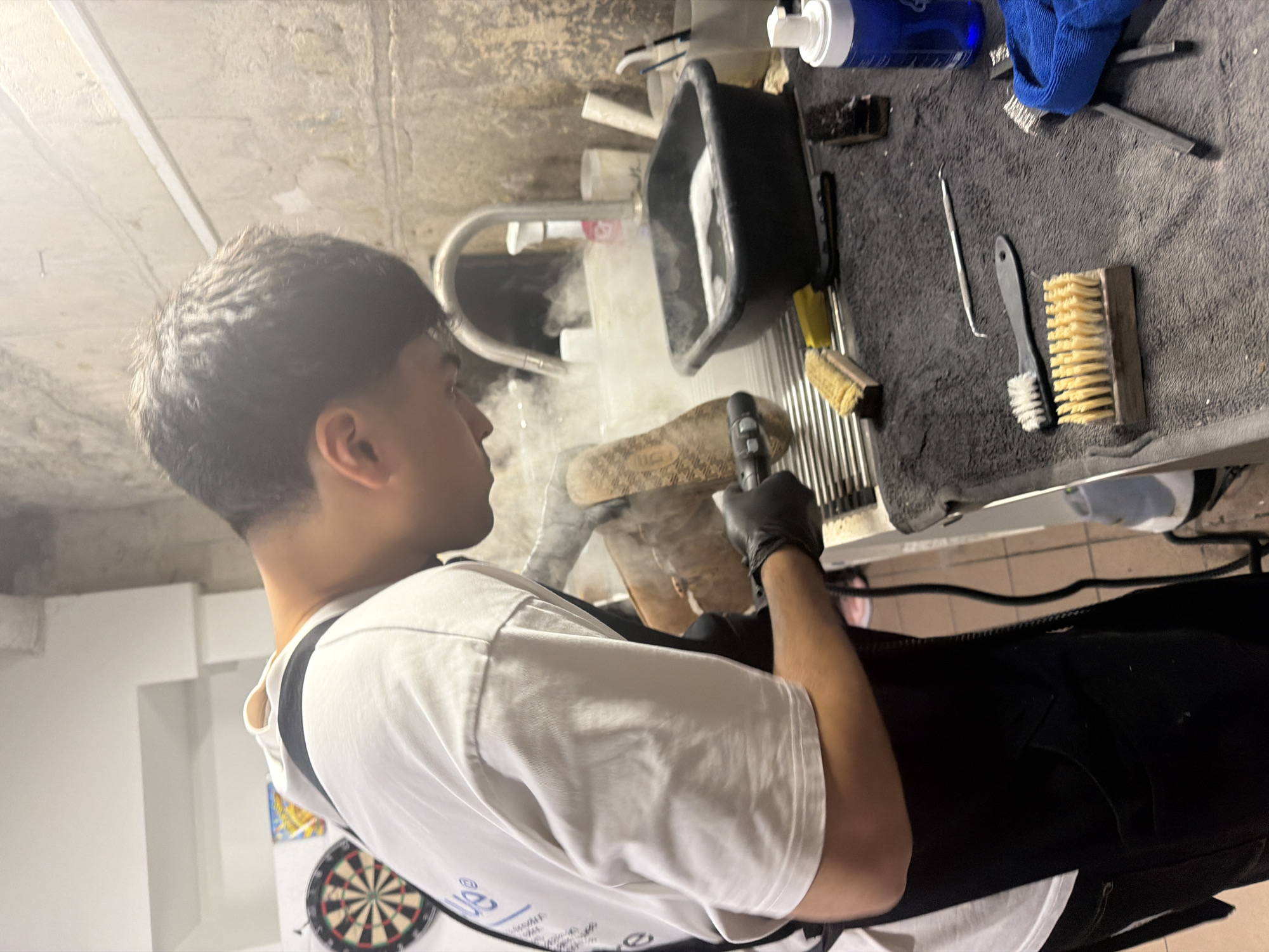 A young man in a white t-shirt and black pants, wearing black gloves, is cleaning a shoe sole with a handheld tool emitting steam. The scene is indoors, possibly a basement or workshop, given the unfinished concrete ceiling and visible support beams. A dartboard is visible on the wall in the background.
On a table to the right, covered with a gray towel, are various cleaning supplies, including brushes, cleaning tools, and a black container with water. The environment appears to be a work area dedicated to shoe cleaning or restoration.
There is no evidence to suggest this image was taken in Paris, France.
