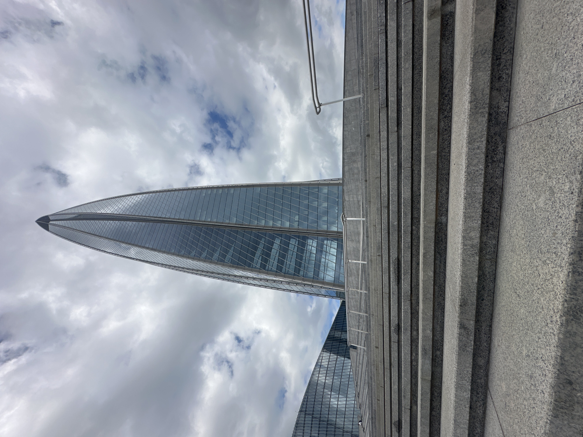 The image shows the Lakhta Center, a 462-meter (1,516 ft) skyscraper in the northwest of Saint Petersburg, Russia. The building is the tallest skyscraper in Russia and Europe and the second-tallest structure in Europe. The skyscraper is shaped like a flame and is covered in glass panels that reflect the sky. The image is taken from a low angle, looking up at the building from a set of granite stairs. The sky is cloudy, with patches of blue sky visible. The stairs are wide and made of large granite slabs, with a metal railing on the right side. In the background, another glass building is visible, likely part of the Lakhta Center complex. No people are visible in the image.