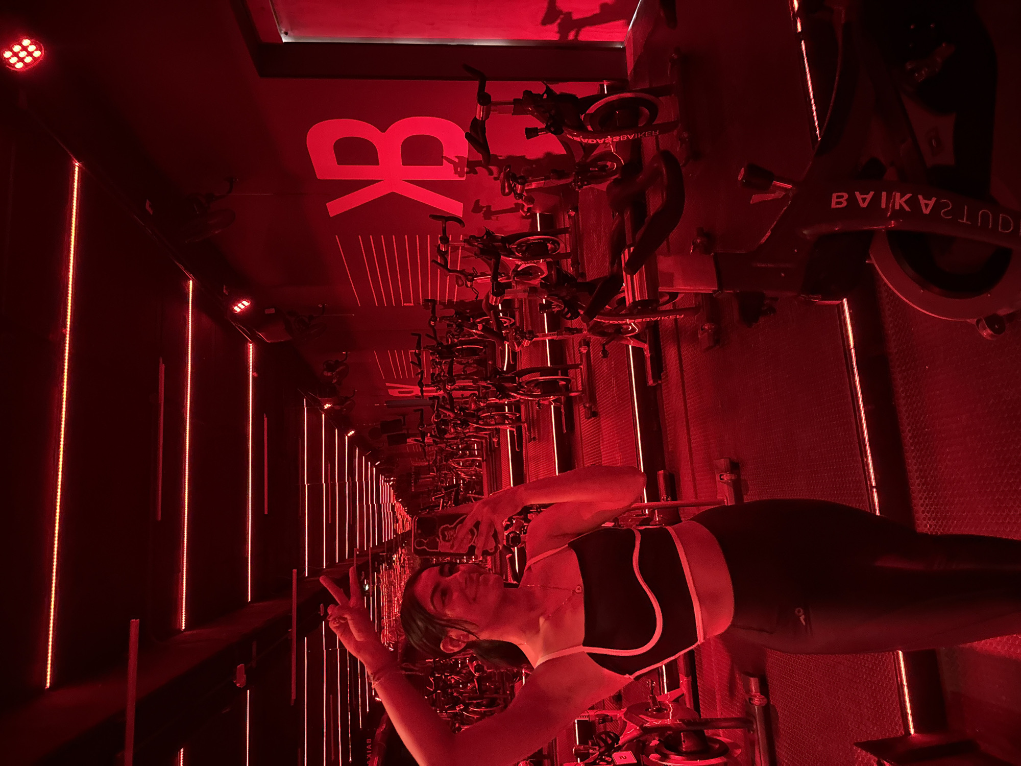 The image depicts a woman in a black and white athletic top and black leggings taking a selfie in a red-lit indoor cycling studio, identified by the backwards "BK" logo and the inverted text "BIKESTUDIO" on the bikes, consistent with a fitness studio in Puebla, Mexico. The studio is lined with stationary bikes and vertical red lights. The woman is holding up two fingers in a peace sign gesture.