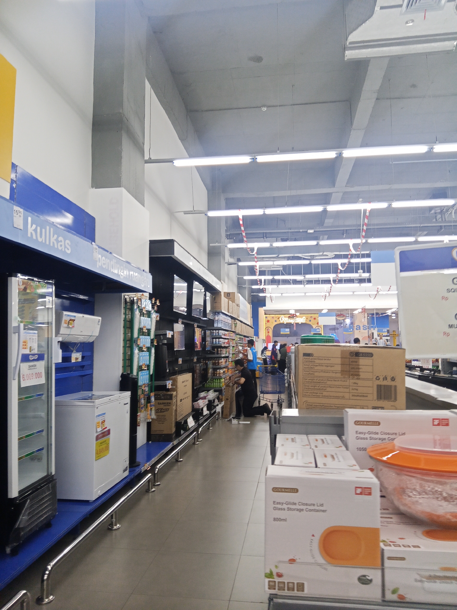 The image captures an aisle inside a supermarket in Lubuklinggau, Indonesia. The aisle features a display of appliances, including refrigerators and air conditioners, on the left. Further down the aisle, shelves are stocked with various household goods, predominantly packaged items. The ceiling is a typical commercial structure with exposed beams and fluorescent lighting. In the background, other shoppers are visible browsing or pushing shopping carts. A security railing runs along the length of the aisle, guiding customer flow. The overall scene depicts a retail environment with an emphasis on home appliances and storage containers.