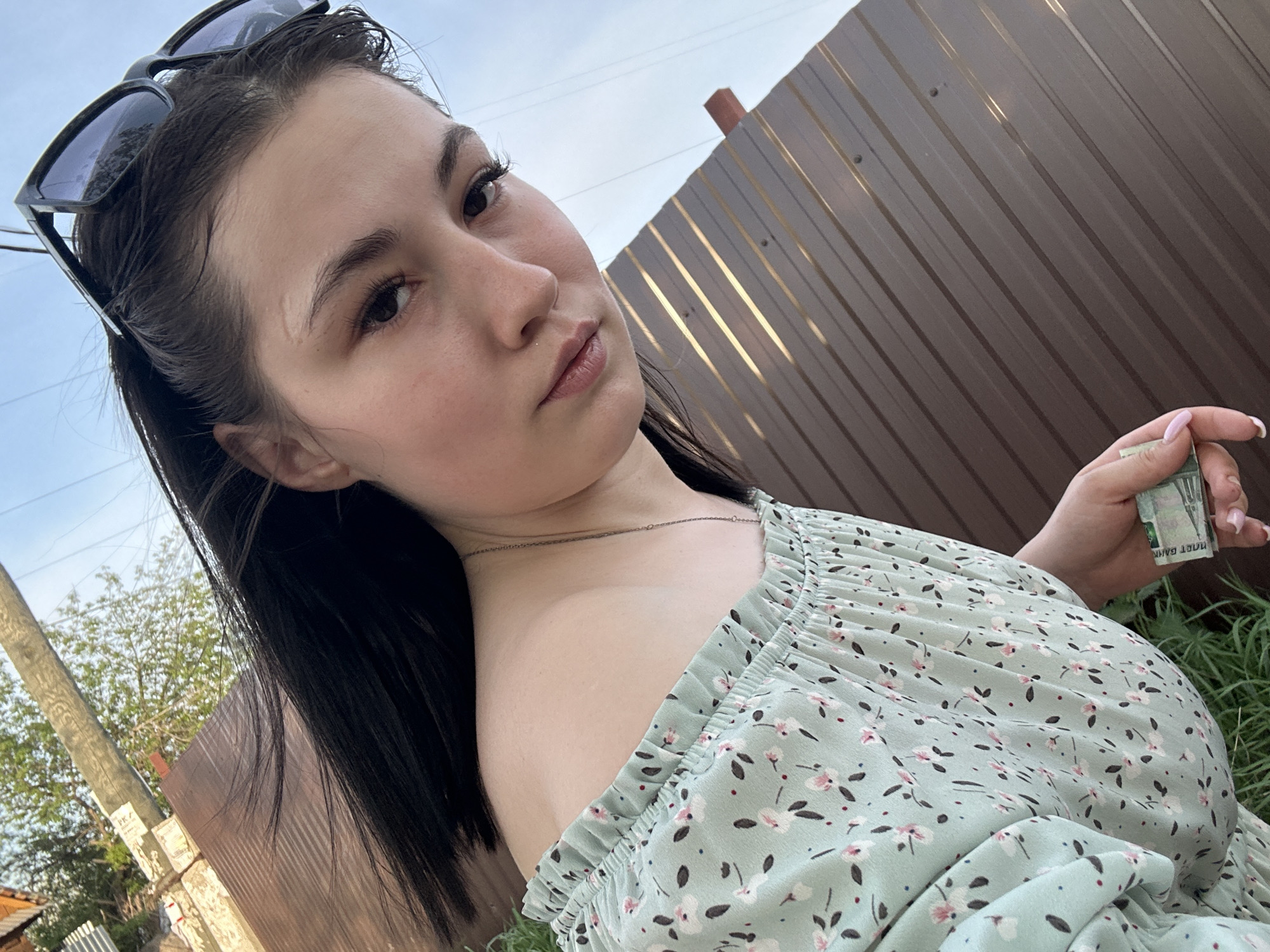 A young woman stands outdoors in Chaikovsky, Russia, holding a wad of Russian currency in her right hand. She is facing the camera with a slightly angled gaze, wearing sunglasses on top of her head and a light green floral dress. The background includes a brown metal fence, a utility pole, and trees. The location appears to be a residential area.