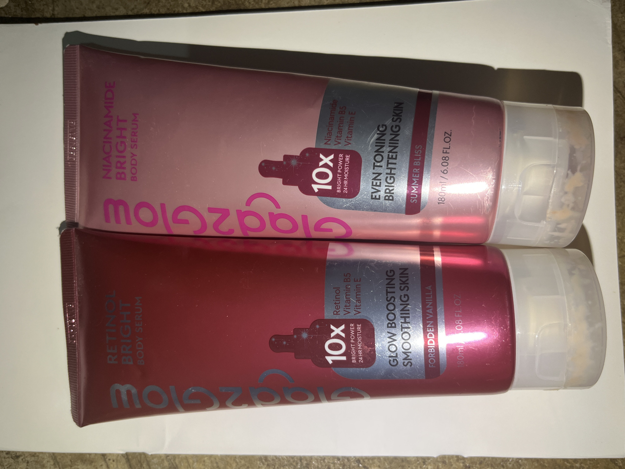 Two tubes of body serum are shown in the image. The tube on the left is pink and labeled "RETINOL BRIGHT BODY SERUM". The tube on the right is a lighter pink and labeled "NIACINAMIDE BRIGHT BODY SERUM". Both tubes have the brand name "Glad2Glow" printed vertically on the left side. Text on the left tube indicates it contains "GLOW BOOSTING SMOOTHING SKIN" and has a scent of "FORBIDDEN VANILLA." The right tube indicates it has "EVEN TONING BRIGHTENING SKIN" and a scent of "SUMMER BLISS." Both tubes show "10x BRIGHT POWER 24 HR MOISTURE" and list key ingredients. The bottom of each tube has a clear cap. The background appears to be a light-colored surface. The image quality is fair. The image was not taken in Meliau Hulu, Indonesia.
