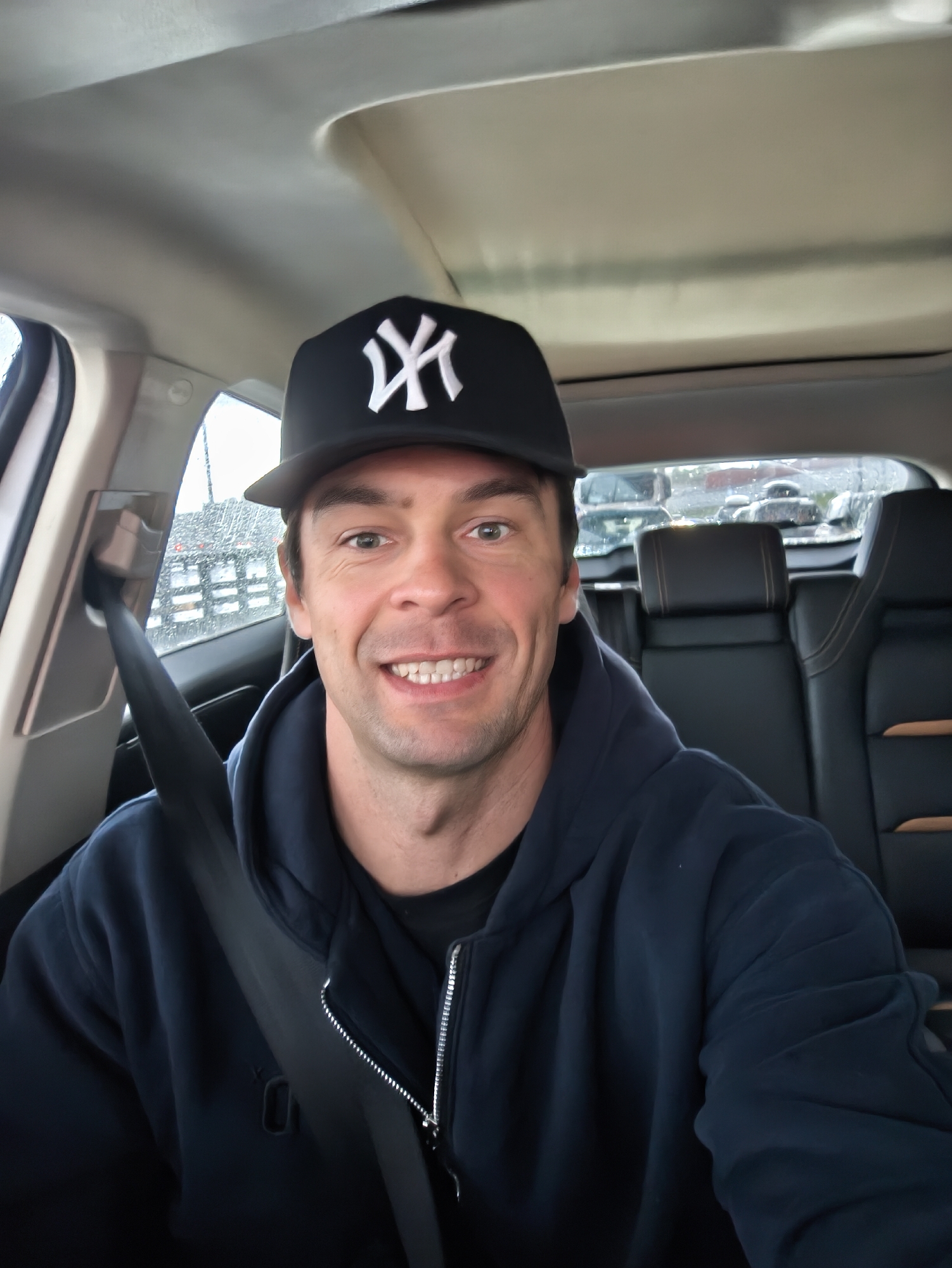 A man wearing a dark blue hoodie and a black baseball cap with a white "NY" logo is seated in a car, wearing a seatbelt. The car's interior includes black leather seats with bronze accents. Outside the car, it appears to be raining, with blurred vehicles visible through the rear window. The location cannot be determined from this image.