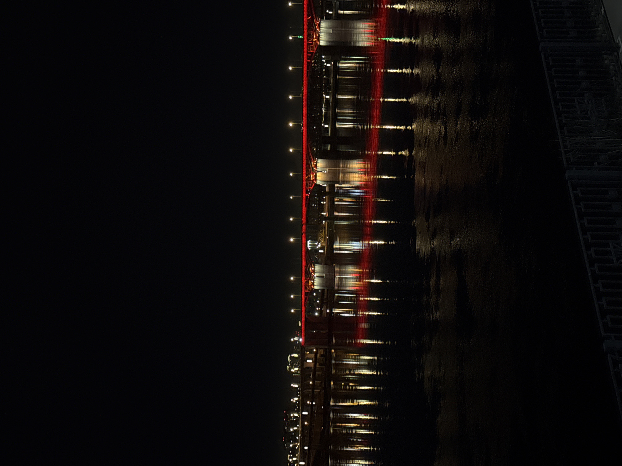 Captured at night, the image presents a sideways view of a bridge reflected in water. The left portion of the frame is nearly completely black, emphasizing the darkness. Along the horizontal center of the image, the bridge's structure is lit with red lights, which are also reflected in the dark water below. The water surface is visibly textured, suggesting movement and imperfect reflection. Additional lights, possibly from the bridge's street lamps, cast straight, elongated reflections on the water. On the right side of the frame, what appears to be a concrete structure with a pattern of rectangular openings is visible. Given the presence of a bridge with red lights reflected in a body of water, this scene strongly suggests the Vinogradovsky Bridge spanning the Yenisey River in Krasnoyarsk, Russia.