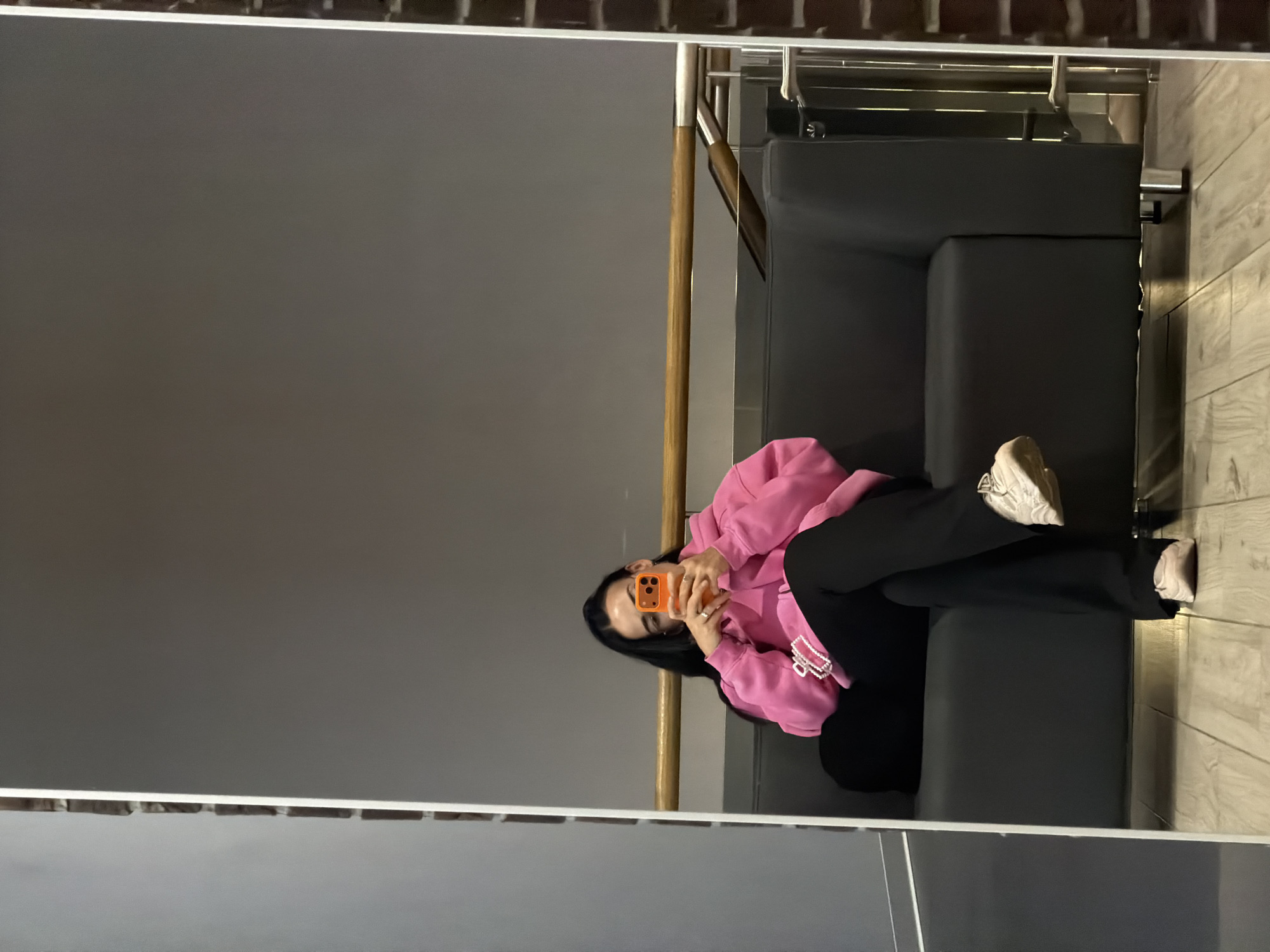 A mirrored image shows a woman sitting on a grey couch. She is wearing a pink hoodie and black pants. She is holding an orange phone in front of her face. The background consists of a grey wall and a wooden floor. The location is a building in Saint Petersburg, Russia.