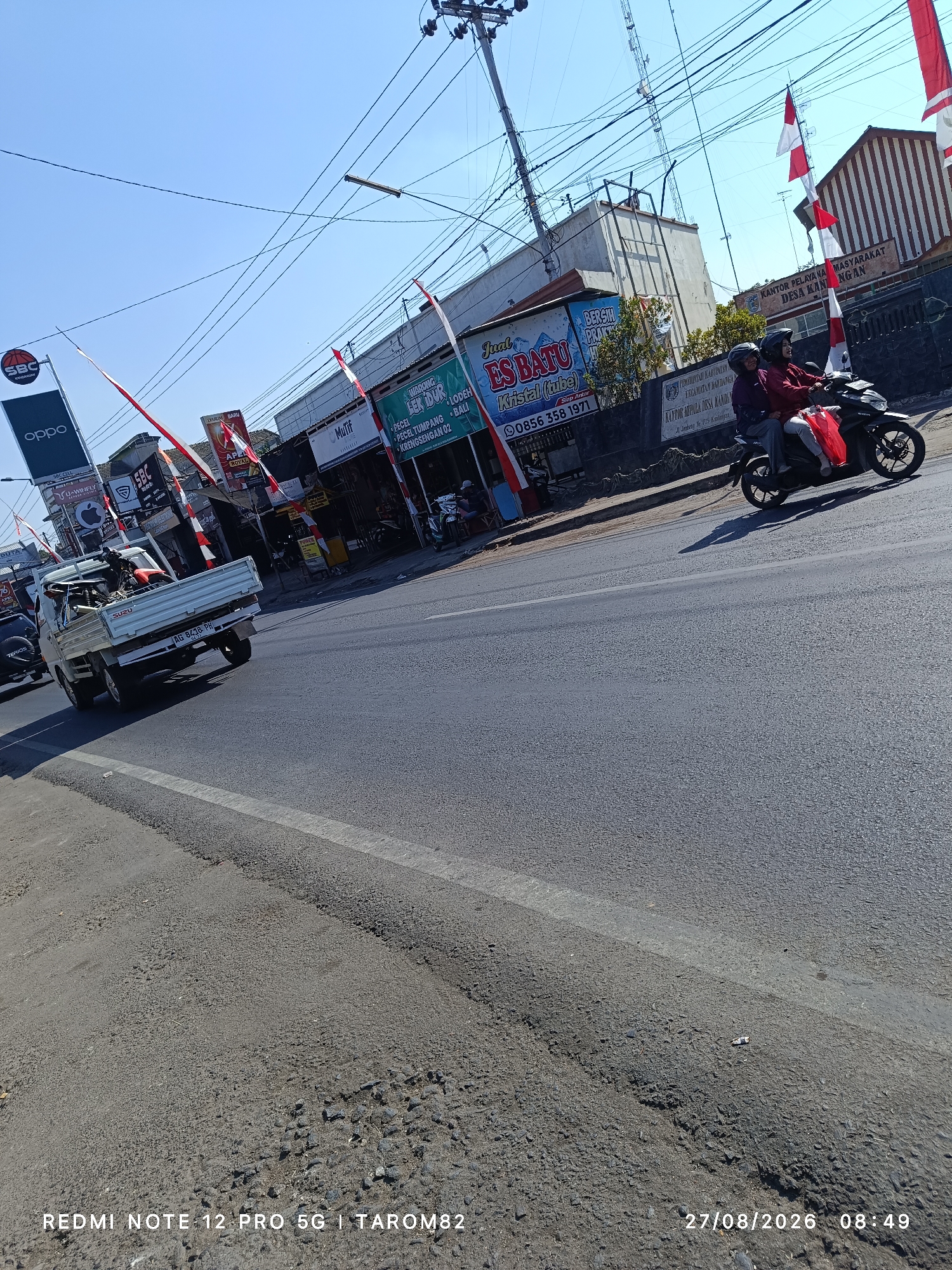 The image depicts a street scene in Kandangan, Indonesia, on August 27, 2026, at 08:49 AM. A white, three-wheeled pickup truck with the license plate "AG 9438 PH" is driving on the right side of the road, carrying various items in its bed, including what appears to be motorcycles. A black motorcycle with two occupants, a driver and a passenger wearing helmets and red jackets, is in front of the truck, also traveling on the right side of the road. On the left side of the road, there are several storefronts with signs, including "OPPO," "SBC CELL," and "WARUNG LEK LODEH." Red and white Indonesian flags are displayed prominently along the street. The road surface shows signs of wear and tear, with visible patches and cracks. The sky is clear and blue, indicating a sunny day. The background features buildings, utility poles, and a large transmission tower.