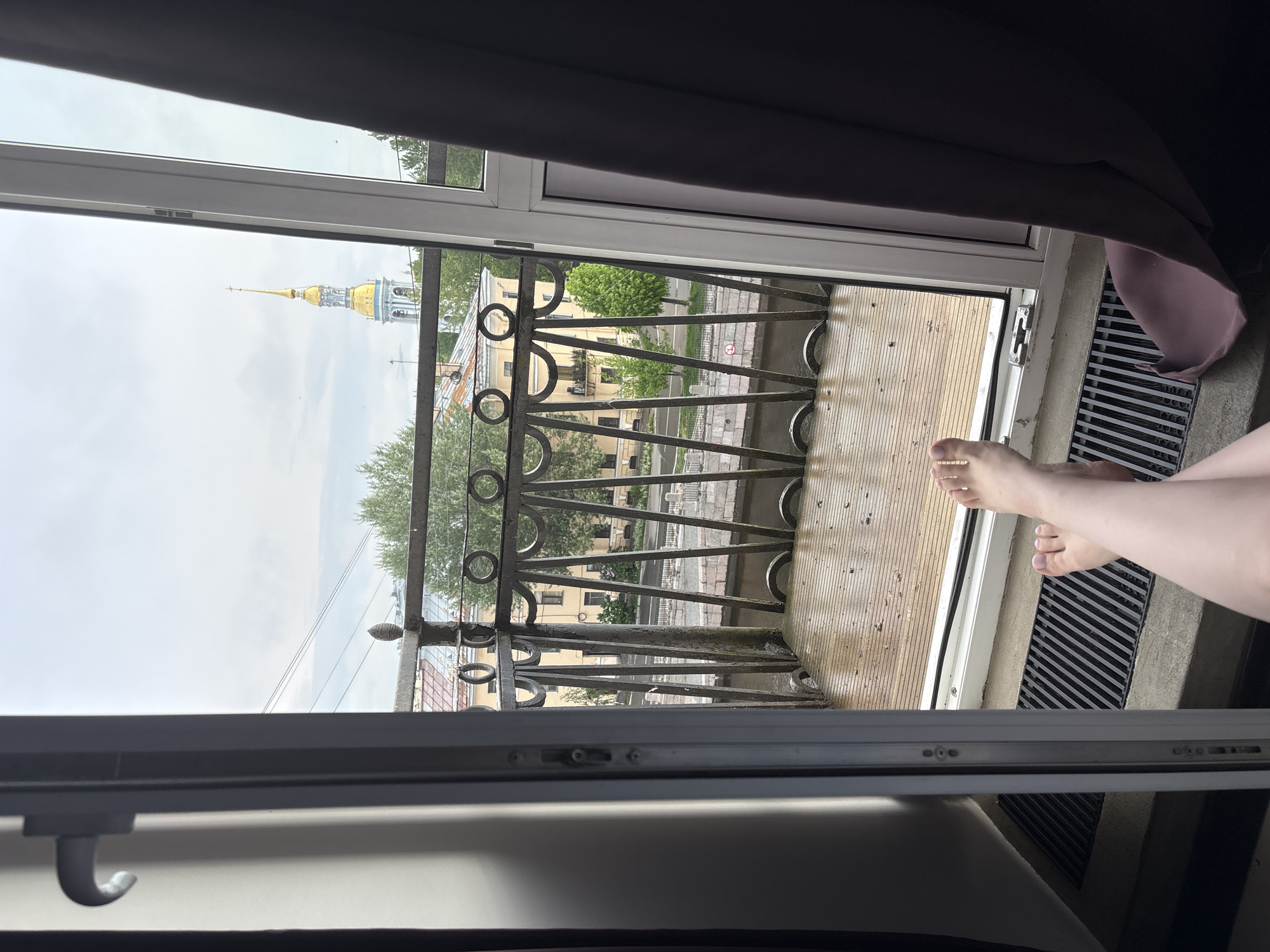 The image shows a view from an open balcony door, revealing an outdoor scene and part of an interior space. The balcony features an ornate, dark metal railing with circular and triangular designs. Through the railing, a street scene is visible, with buildings, trees, and a portion of what appears to be a gilded spire in the distance.
Inside, a portion of bare legs and feet are visible near the bottom right of the frame. The floor of the balcony is light-colored and appears to be wet or damp. The interior space includes part of a closed window, dark curtains, and a light-colored surface.
The gilded spire in the distance is the main spire of St. Nicholas Naval Cathedral. This places the location in the city of Saint Petersburg, Russia.
