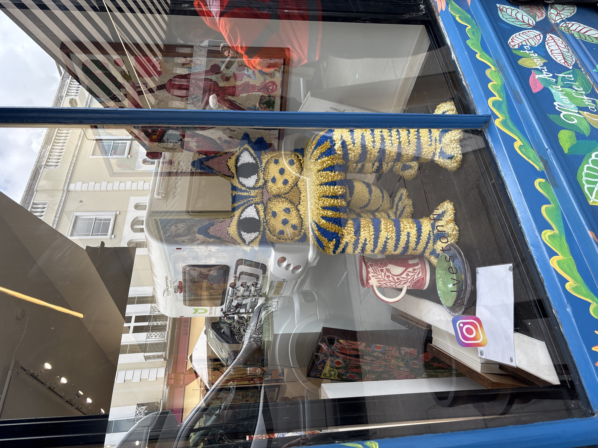 The image depicts a shop window display. Inside the window, there is a yellow and blue striped stuffed animal resembling a tiger, various books, and a mug. The shopfront is painted blue and features floral and leaf designs in green, pink, and white. Text reading "Maria Bonneto" can be seen on the painted shopfront. An Instagram logo is stuck to the glass. The reflection in the window shows a building with windows, awnings, and an awning with stripes. The name "Alveston" is present on a sticker in the shop window. Based on this clue, the image could have been taken near Alveston, Warwickshire, in the United Kingdom.