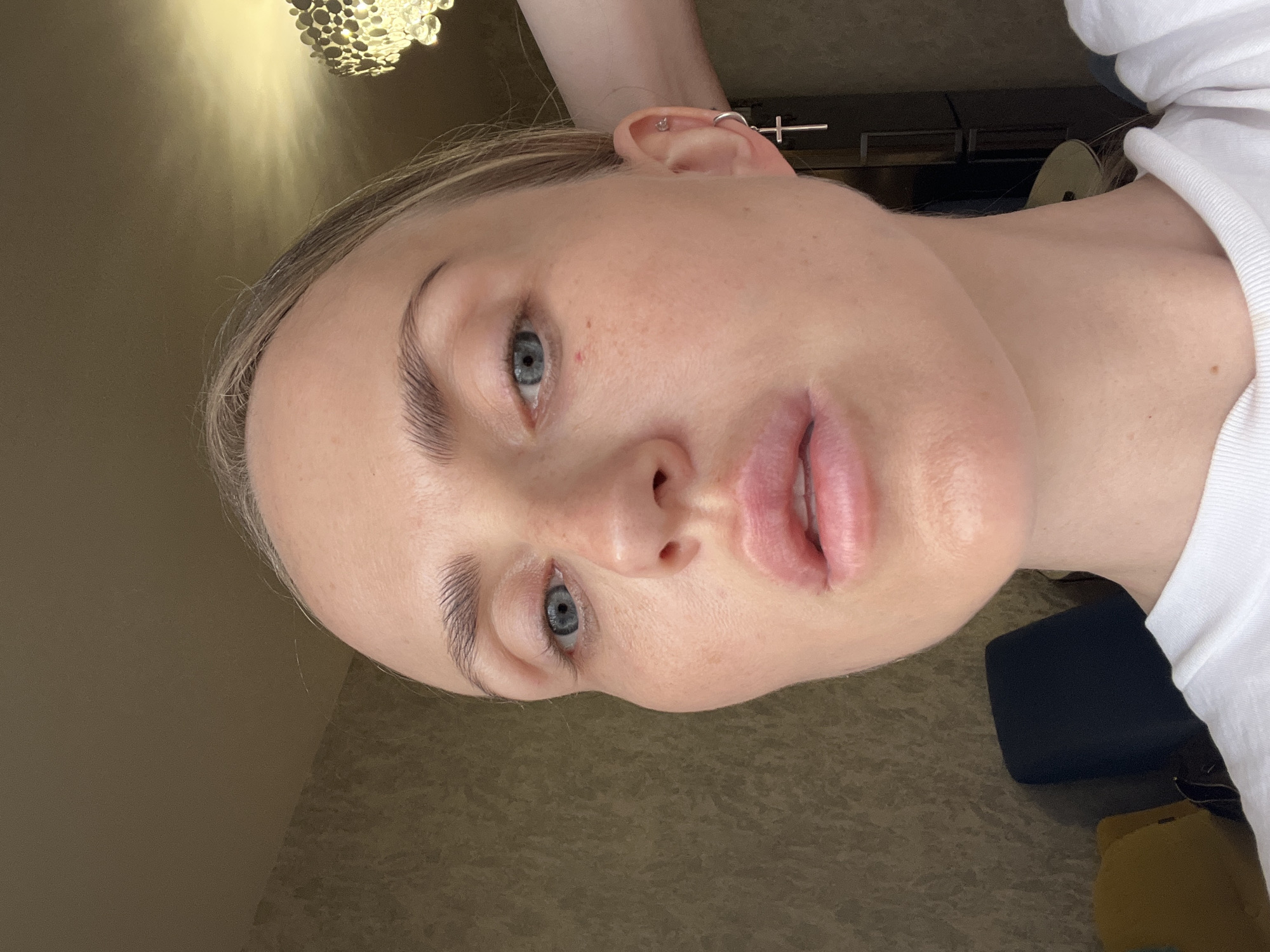 A close-up, slightly angled, waist-up portrait of a Caucasian woman with fair skin and blonde hair pulled back. She is looking slightly to her left with a neutral expression. Her makeup is minimal, highlighting her natural features. She is wearing a white t-shirt and a silver cross earring in her left ear. The background appears to be an indoor setting with textured wallpaper and a modern chandelier visible on the right side. There is a chair and a hint of a yellow object in the lower left corner. The lighting suggests it is daytime, with natural light coming from the right.