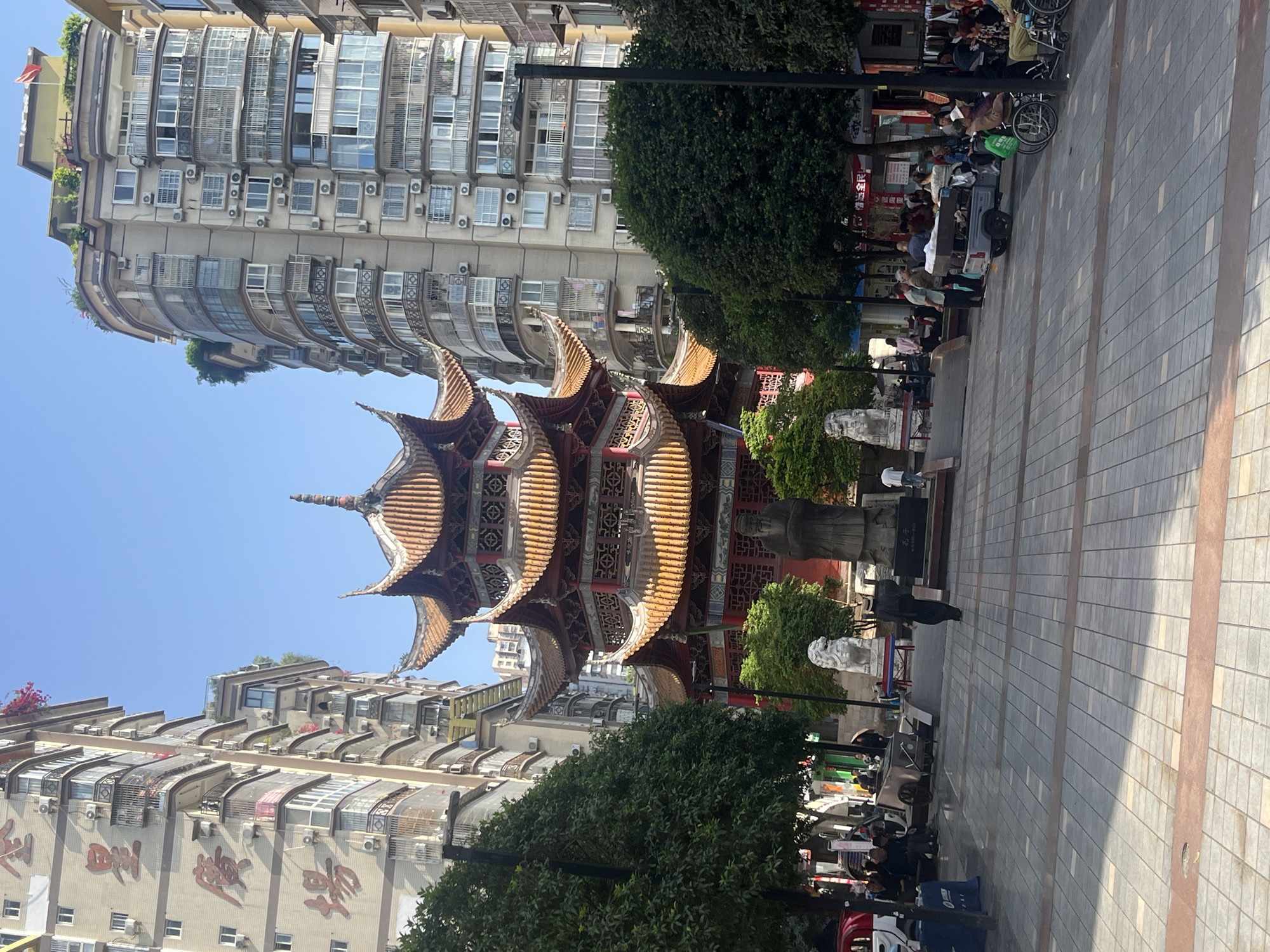 This image captures a vibrant urban scene in Jiangjin, China, showcasing a blend of traditional Chinese architecture and modern city life.
At the center is a magnificent, multi-tiered pagoda, distinguished by its intricate, curving roofs tiled in orange and brown, adorned with ornate red and gold decorations. This traditional structure is flanked by tall, modern residential or commercial buildings. These contemporary buildings are characterized by numerous windows, balconies (some enclosed), and visible air conditioning units. The building on the left prominently features large, red Chinese characters on its facade.
The ground is a wide, paved plaza with grey and brown rectangular tiles, suggesting a public pedestrian area. Numerous people are visible throughout the plaza, indicating a lively atmosphere. Many individuals are seated on benches or chairs along the right side of the plaza, with several wheelchairs suggesting a gathering place for community members, possibly including the elderly. A small, canopy-covered cart or vehicle is also present. Statues, including a white stone lion and another darker figure, are positioned along the walkway, near the base of the pagoda.
The clear blue sky and bright lighting indicate a sunny day, likely late morning or early afternoon. The scene portrays a bustling public space where traditional culture coexists with the functional aspects of a modern Chinese city.