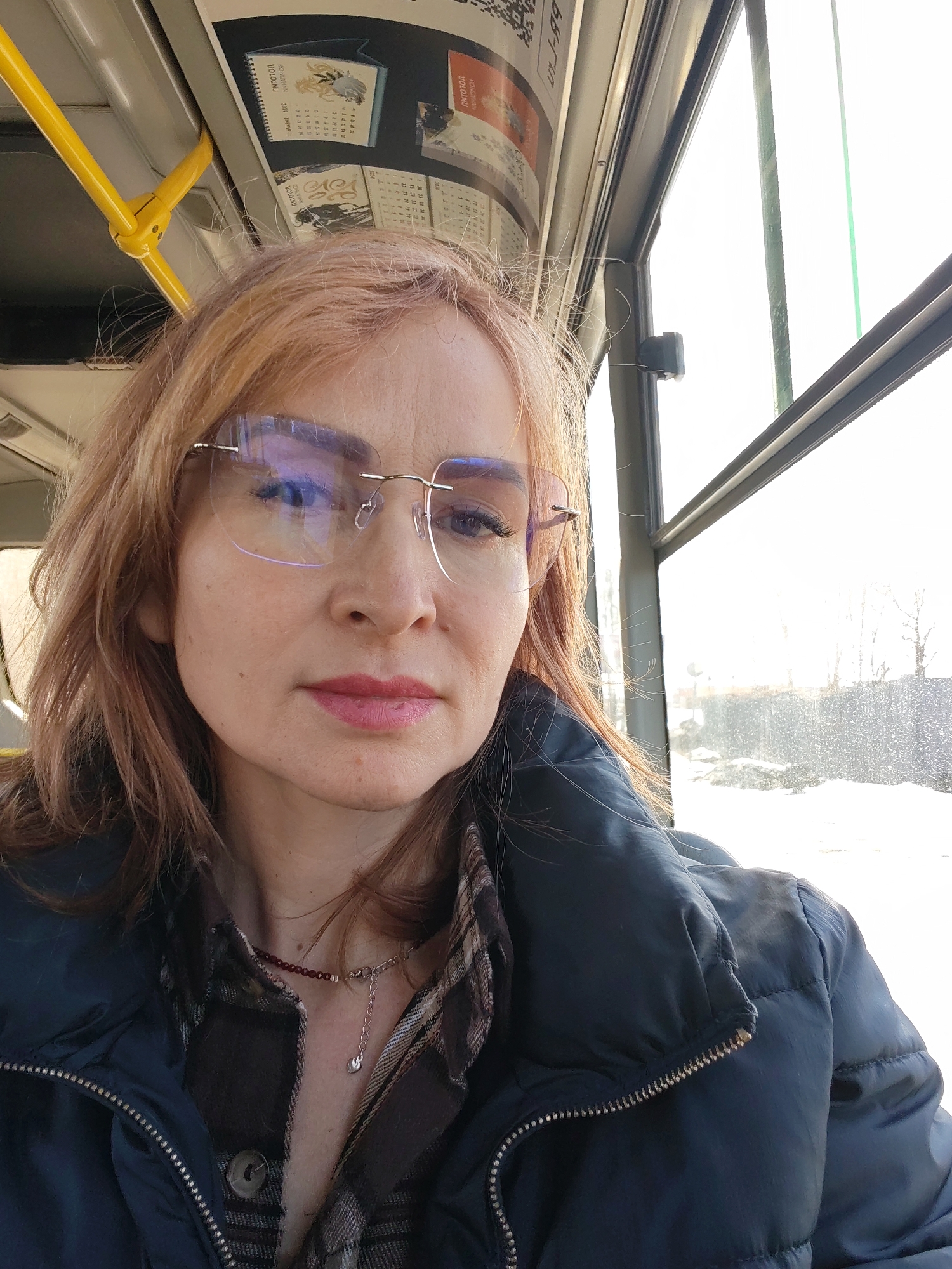The image depicts a Caucasian woman inside a public transport vehicle, likely a bus, in Chaikovsky, Russia. She is shown in a selfie-style composition, looking directly at the camera with a neutral expression. She has light reddish-brown hair and wears rimless eyeglasses, the lenses of which reflect purplish-blue light. Her facial makeup includes light pink lipstick. She is dressed in a dark blue puffer jacket, unzipped to reveal a plaid shirt in shades of brown and beige. She wears two necklaces: a short, dark red beaded strand and a longer silver chain with a small, teardrop-shaped pendant.
To the subject's right, a vehicle window reveals a bright, snowy exterior landscape, including bare trees and a dark fence or barrier. Above the woman, a yellow overhead grab bar and a rack containing advertisements and calendars with Cyrillic text are visible, consistent with a Russian public bus interior.