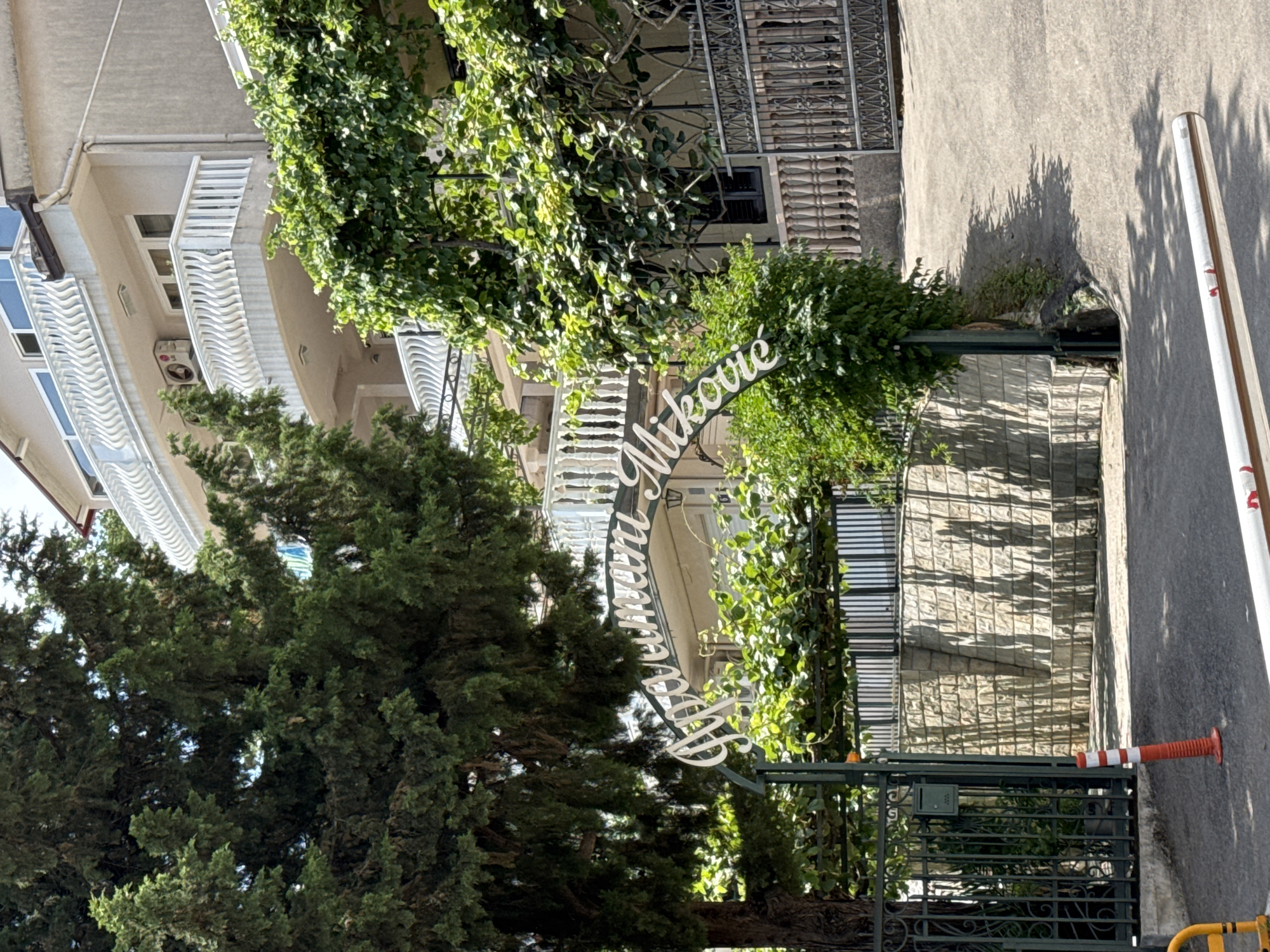 The image shows an outdoor scene in what appears to be a residential area or a complex. A building with white balconies and a light-colored facade is visible on the left. Lush green trees and climbing plants surround the area, some of which are growing over an ornate dark green metal gate. An archway is formed by a vine-covered pole, with white cursive text on it. The text is partially obscured by foliage and the angle of the image but appears to read "Hotel Miković" or similar, with "Hotel" written vertically and "Miković" horizontally.
The ground surface is paved with concrete and stones, and a dark asphalt road is visible to the right. A white and red striped traffic cone is placed on the asphalt. The lighting suggests it is daytime, with shadows cast on the ground, indicating a sunny day. The overall impression is one of a well-maintained property with a welcoming entrance, possibly a hotel given the text. The scene is static, with no people or overt activity. The location cues, such as the architectural style and the presence of Mediterranean vegetation, along with the context provided, point to Budva, Montenegro.