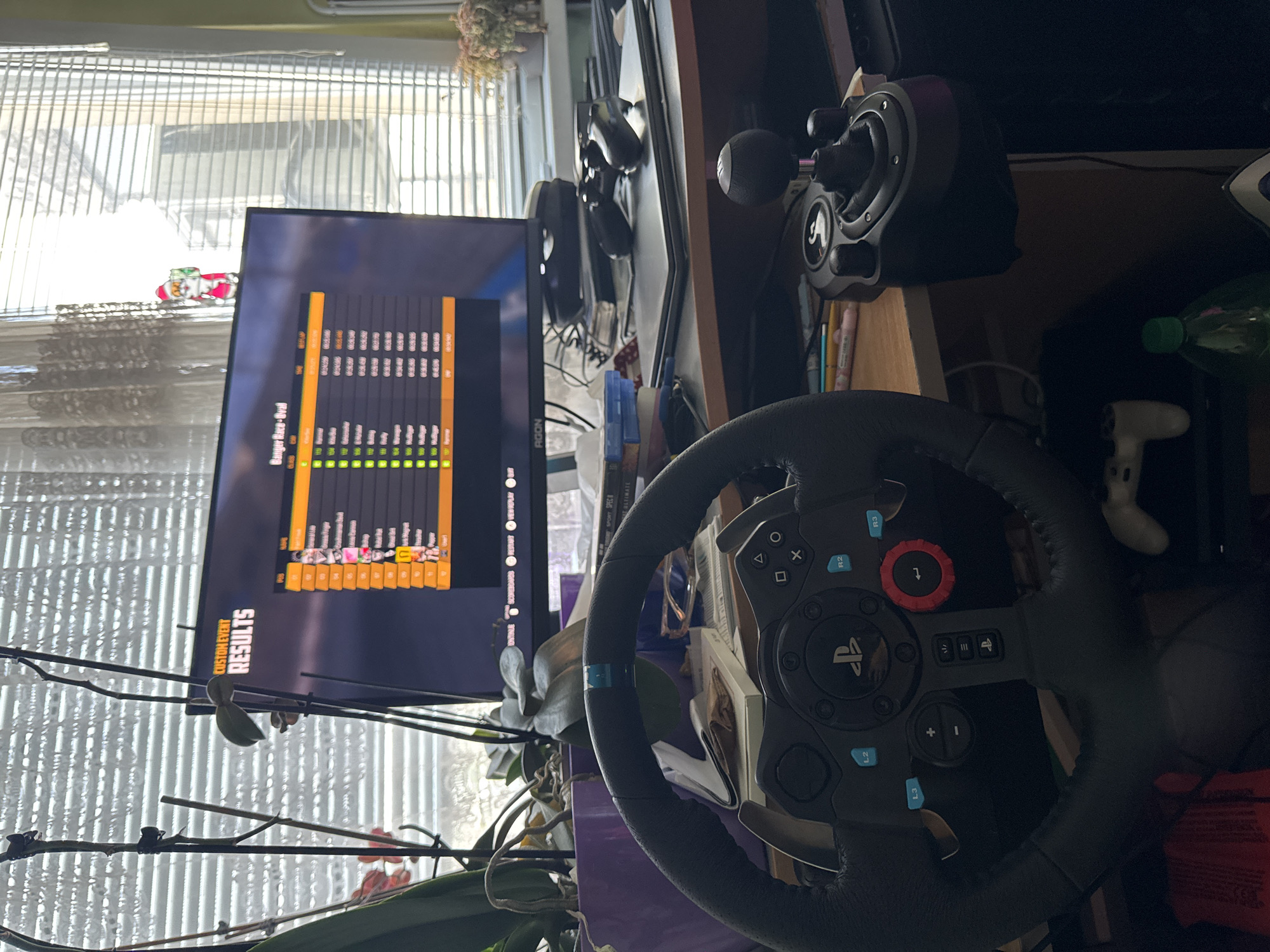 The image shows a racing setup in an indoor environment. A monitor displays a "Custom Event Results" screen for a "Banger Race-Oval" on what looks like an AGON monitor, alongside a Logitech G923 racing wheel with Playstation controls and gear shifter. The scene includes personal items, such as a pen holder and headphones. The overall setting appears to be a casual gaming or entertainment area. The environment includes a window with blinds.