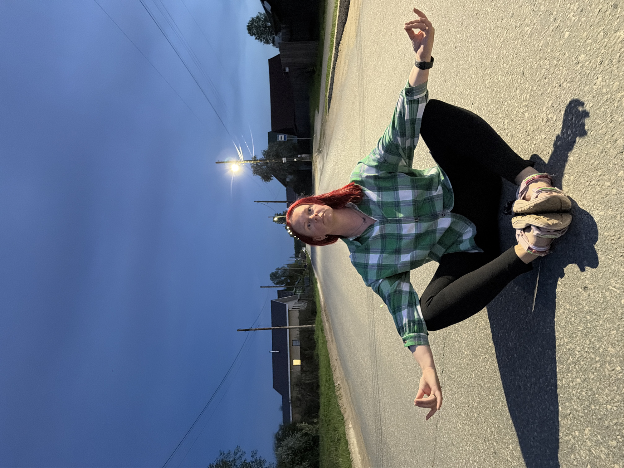 A woman with red hair sits cross-legged on an asphalt road in Chaikovsky, Russia. She wears a green and white plaid shirt, black leggings, and sandals. One arm is raised with fingers extended. The sky is a darkening blue. Houses and utility poles line the road. A street lamp illuminates the scene. Her shadow stretches out on the road.