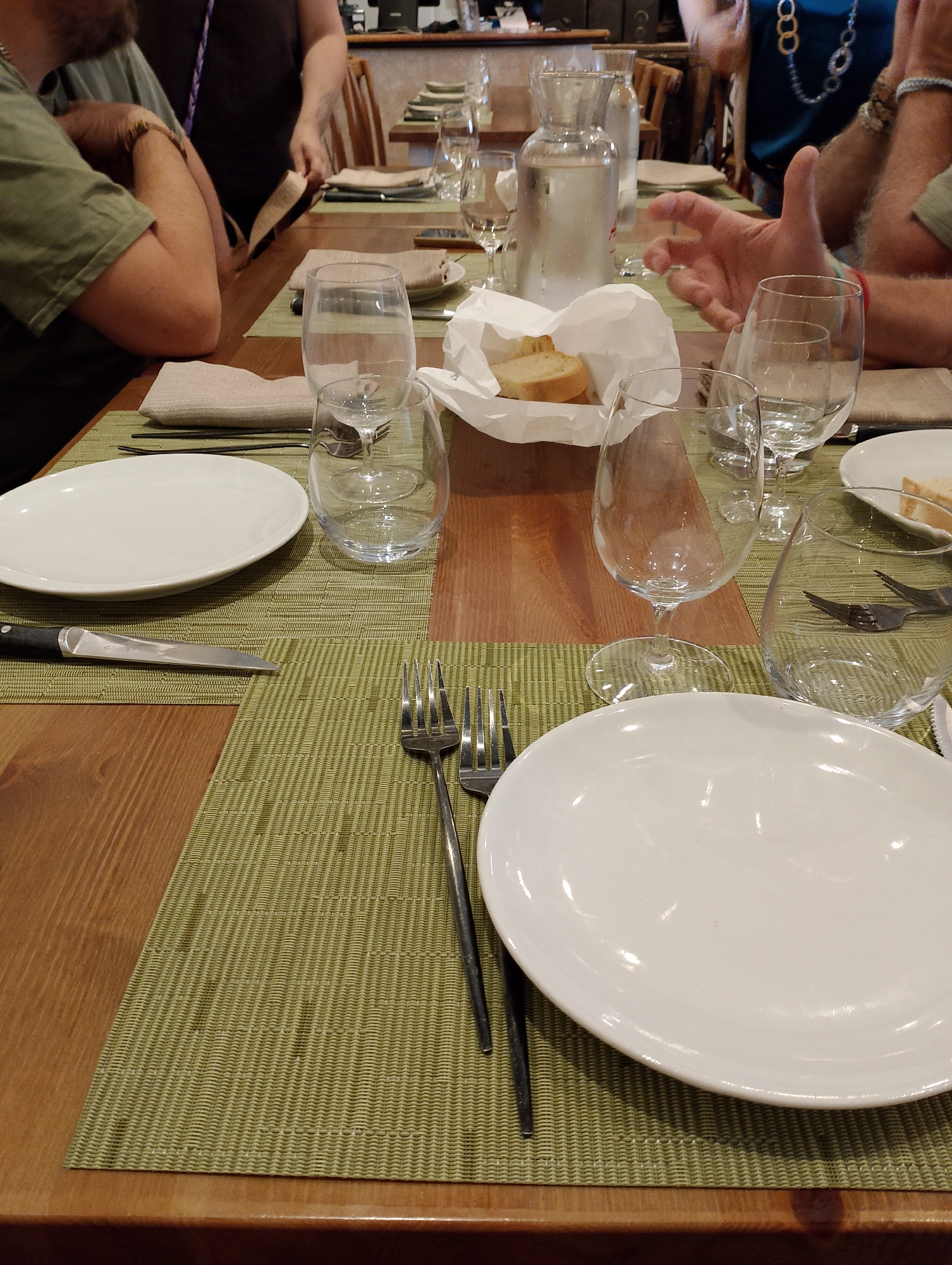 This is a close-up, eye-level, medium shot of a wooden table set for a meal at what appears to be a restaurant, with multiple place settings. The table is covered with light green woven placemats. On the left side of the frame, a white dinner plate is positioned on a placemat, with a knife placed to its left. Further to the left, a person wearing a green t-shirt and a partial view of their arm and hand are visible. They are wearing a bracelet. In the background, other people are partially visible. To the right of the plate, a clear wine glass is empty. Behind it, another clear glass is visible, with forks inside. Further to the right, another white plate with bread and a paper napkin is set. A carafe filled with water and other glasses are also visible. The overall setting suggests a communal dining experience. The restaurant is located in the city of Torri in Sabina, Italy.