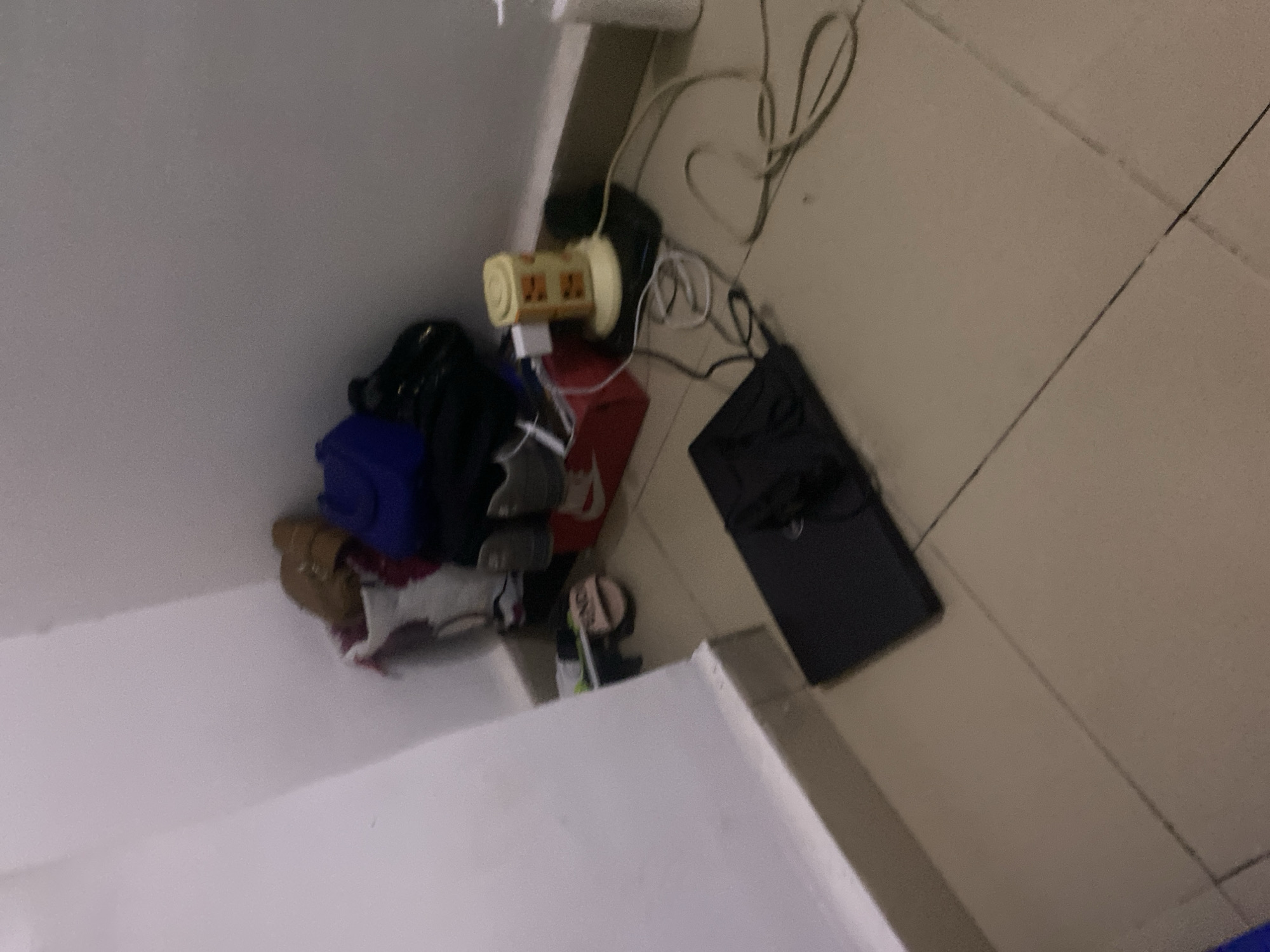 The image depicts a corner of a room, likely a bedroom or storage area, with a tiled floor. In the foreground, a black laptop is open on the floor, surrounded by various electrical cords. To the right of the laptop, a yellow cylindrical object with multiple outlets, presumably a surge protector or power strip, is plugged into a black base. Wires are connected to this device and extend across the floor. Further to the right, a white bottle, possibly a lotion dispenser or soap, stands near the wall.
In the background, a collection of items is piled near the corner. This includes a red shoe box with a white Nike logo, and several pairs of shoes, including athletic shoes and what appear to be casual slides. Bags, one a dark blue, are also stacked amongst the shoes. The wall in the background is a neutral color, and a white partition or wall extends from the left side of the image. The overall impression is one of clutter and a functional, lived-in space.
The specific location of Howayi, Nigeria, is not determinable from the visual evidence in the image itself. This scene is common in many urban and residential settings and does not contain any unique identifiers tied to a specific geographic location within Nigeria.