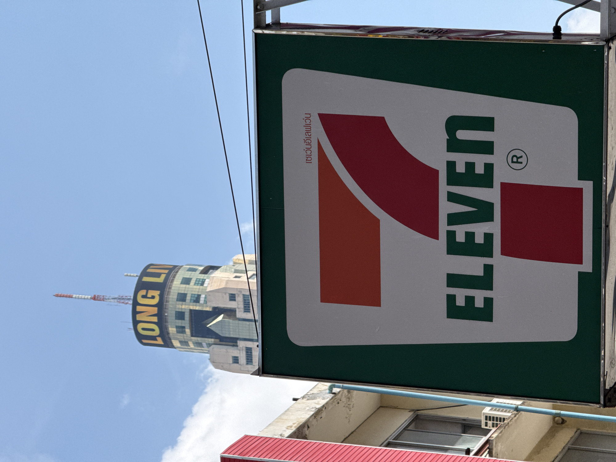 The image shows a 7-Eleven sign in the foreground, with a tall building labeled "LONG LI" visible in the background. The sign is the standard 7-Eleven logo with its recognizable color scheme. The building in the background is a cylindrical structure with the "LONG LI" sign displayed prominently near the top. The sky is visible, suggesting an outdoor setting. The location is likely in Bangkok, Thailand, based on the presence of the Long Li building which is known to be located in the city, as well as the fact that many 7-Eleven stores are in Thailand. The rest of the surrounding buildings are low-rise structures.