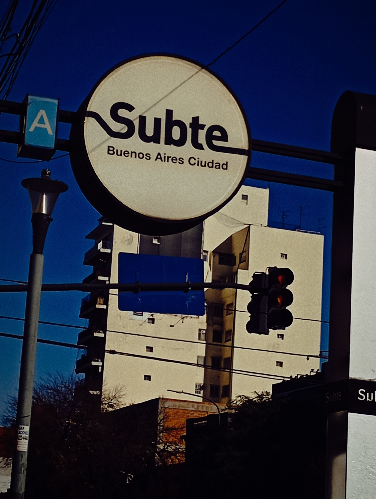 The image depicts a street scene in Buenos Aires, Argentina. The primary subject is a circular sign for the "Subte" (subway), specifically indicating Line A. "Buenos Aires Ciudad" is written on the bottom part of the sign. The sign is mounted on a black metal pole. To the left is a light post.
Behind the sign and pole is a tall, multi-story building with a mix of light-colored exterior walls. A blue rectangle obstructs the face of the building. A traffic light with red lights showing is on the building.
Electrical wires run across the top of the frame against a deep blue sky. Dark silhouettes of trees and foliage are visible in the lower portion of the image. A white sign with the words "Subte" are also visible on the right side of the frame.