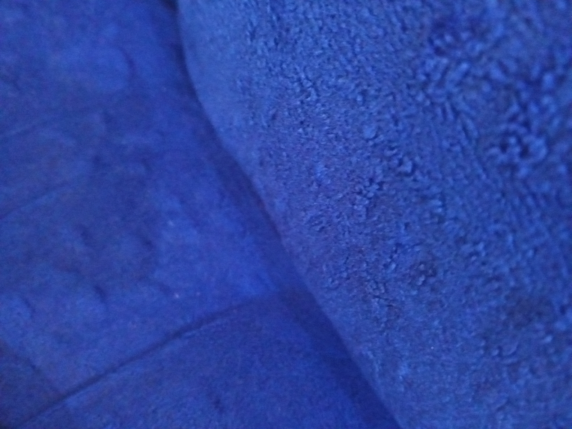 The image is a close-up, abstract shot of a blue, textured fabric, possibly a towel or blanket. The texture appears plush and soft, with a slightly uneven surface. The lighting casts subtle shadows, highlighting the fabric's folds and contours. There are no discernible objects, people, or environmental features in the image to indicate a specific location in Nyabe, Nigeria.