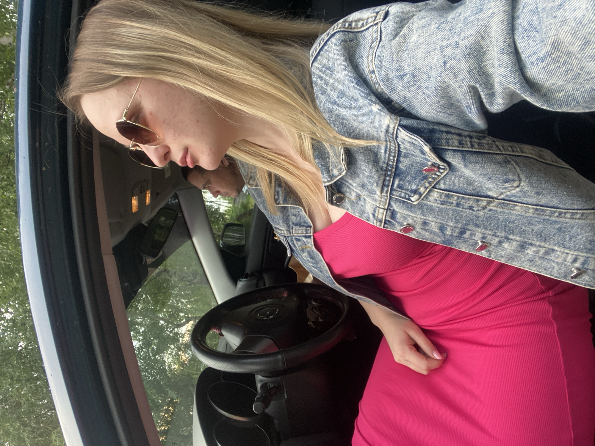 The image shows a young woman with blonde hair and sunglasses sitting in the driver's seat of a car. She is wearing a pink ribbed dress and a denim jacket. Her left hand is resting on her thigh. In the background, a man is visible in the passenger seat. The car's interior is dark, with a steering wheel and dashboard visible. Through the car windows, trees and foliage can be seen, suggesting an outdoor setting, possibly during the daytime given the natural light. The overall mood appears casual and relaxed. The image is taken from a close-up perspective, focusing on the woman and the car's interior.