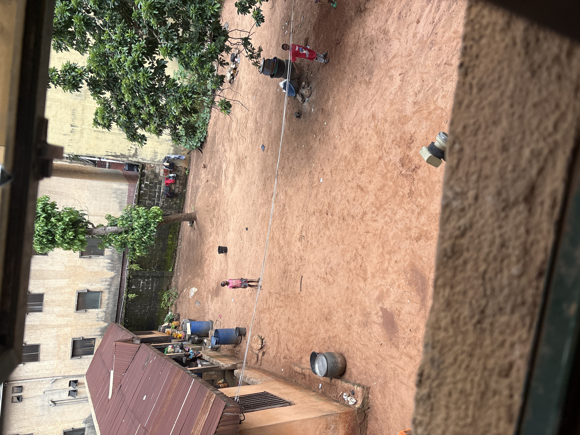 The image depicts an outdoor courtyard area in a residential setting in Onitsha, Nigeria. The ground is predominantly red earth. In the foreground, a weathered, wooden structure with a rusty, corrugated metal roof occupies the left side of the frame. Adjacent to this structure, two large, dark blue barrels and a grey metallic pot are visible. Further into the courtyard, a young child in a pink shirt and patterned shorts stands holding a stick. In the mid-ground, a large tree with lush green foliage stands near a wall and some laundry is hung out to dry on a line. To the right, a cluster of dark, stacked containers, possibly for water storage, is present. Another child, wearing a red t-shirt and grey trousers, is standing near these containers. In the background, multi-story buildings with neutral-colored facades and numerous windows are visible. The overall scene suggests a typical day in a densely populated urban environment.