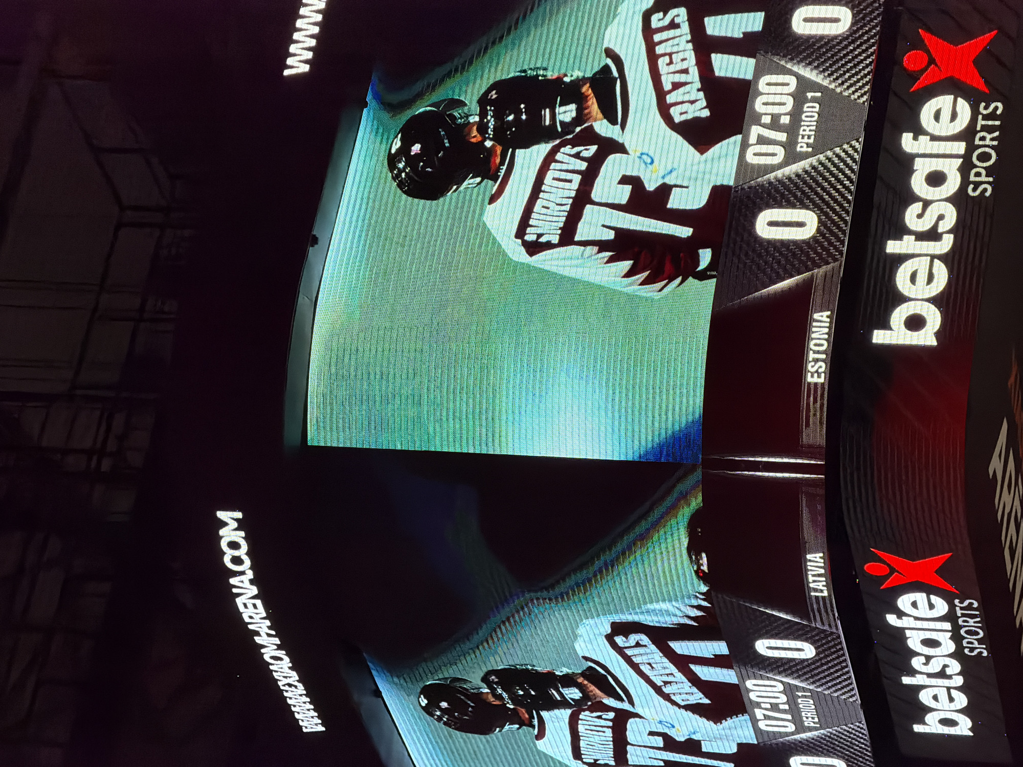The image shows a digital scoreboard at the Olympic Sports Center Arena in Riga, Latvia. The scoreboard displays information about a hockey game. On the left side of the scoreboard, the word "LATVIA" is visible, and on the right side, the word "ESTONIA" is visible. Above the scoreboard, a large screen displays two hockey players in uniform, with their names and jersey numbers visible. The player on the left has "SMIRNOVS" and "73" on his jersey. The player on the right has "RAZGALS" on his jersey. Both players are wearing helmets and appear to be standing on the ice. Below the scoreboard, there is a large advertisement for "betsafe SPORTS". The overall environment appears to be a sports arena, likely during a hockey game or event.