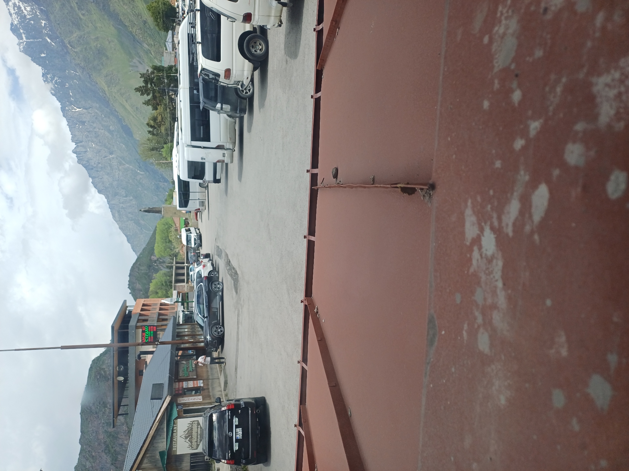 A wide-angle, eye-level, exterior, daytime, full shot depicts a street in a mountainous region. The foreground is dominated by a rusty, corrugated metal roof with visible seams and some white speckling. In the midground, a paved parking lot is filled with various vehicles, including a black SUV parked on the left, a dark SUV in the center, and several white vans and minibuses parked in rows. A person is standing near the black SUV. Beyond the parking lot, a street runs along a row of buildings. The closest building, on the left, is a long, single-story structure with a dark roof, featuring a sign that reads "RESTAURANT". To its right, a multi-story building with a modern design and an orange facade is visible, with a neon sign that appears to say "CHAO". In the background, a towering, snow-capped mountain range stretches across the horizon under a cloudy sky. The overall scene conveys a sense of a remote, scenic location, likely a tourist destination, with vehicles suggesting travel and accommodation services.