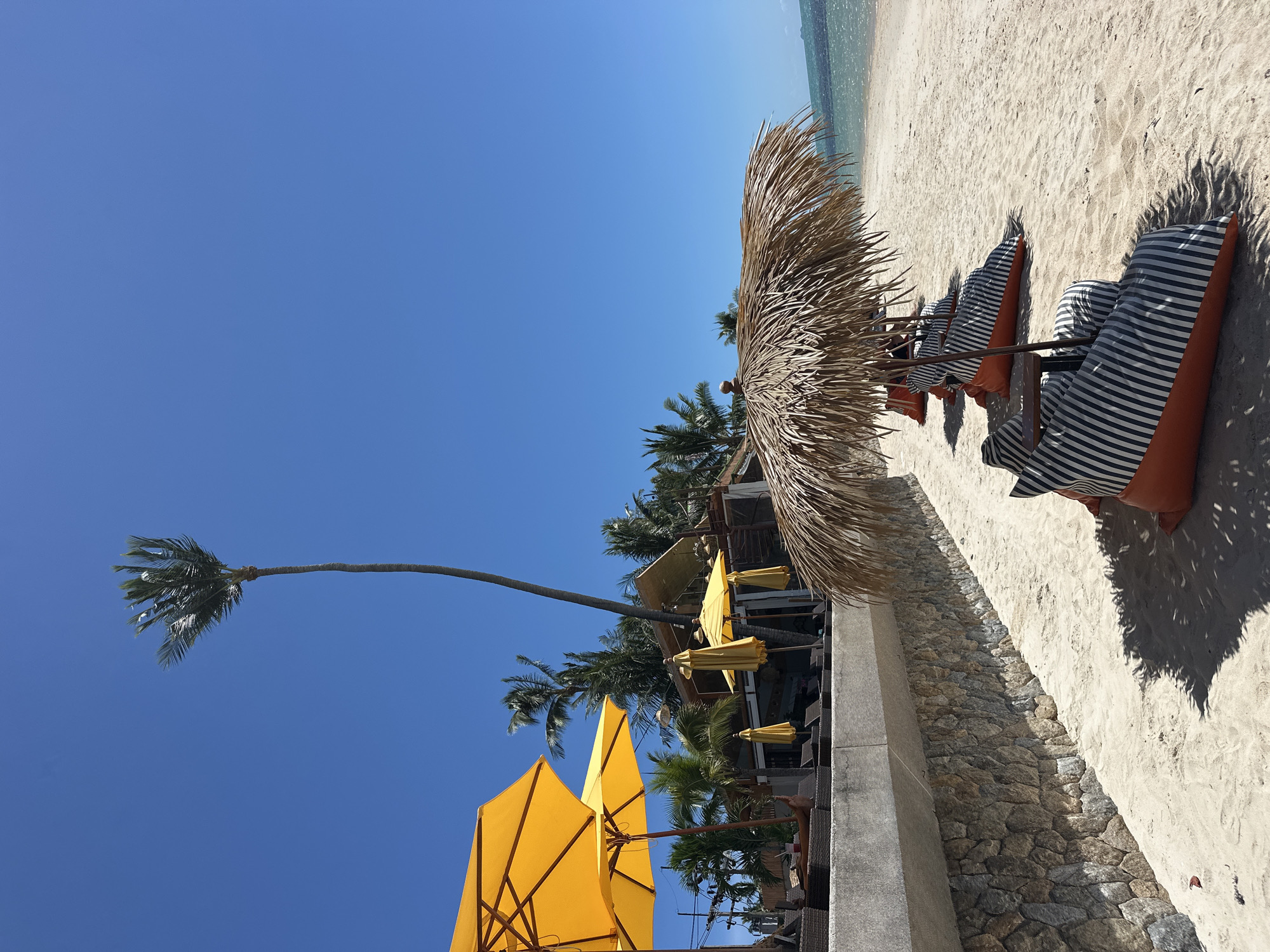 This is a sunlit, eye-level shot of a tranquil beach scene in Baan Tai, Thailand. The image is dominated by a clear, bright blue sky. In the foreground, a row of orange beanbag chairs with black and white striped covers are neatly arranged on a sandy beach. Above them, a thatched umbrella provides shade. To the left, a stone wall with visible mortar runs horizontally across the frame, separating the beach from a slightly elevated area with lounge chairs and yellow umbrellas. Behind the lounge area, palm trees and a building with a wooden roof are visible. The ocean is visible in the distance on the right side of the image, with gentle waves lapping the shore. The overall impression is one of a relaxed, tropical vacation setting.
