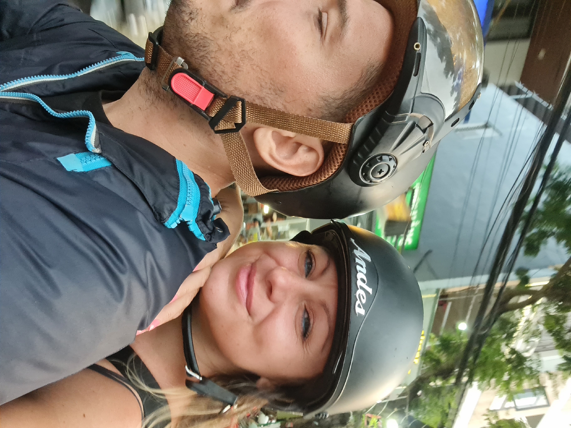 This selfie-style photograph, rotated 90 degrees clockwise, captures two individuals wearing helmets, likely in an urban setting in Đà Nẵng, Vietnam.
In the foreground, a woman with blonde hair and blue eyes is smiling softly towards the camera. She is wearing a matte black helmet with the word "Andes" inscribed in white script on the side. A black strap from her helmet is fastened under her chin, and a black bra strap is visible on her shoulder, beneath what appears to be a dark top. Her fingernails are painted a light pink.
Behind and slightly above her, a man's face is partially visible in profile. He has short dark hair and stubble. He is wearing a dark blue jacket with distinctive light blue zippers and a dark helmet with a clear visor. His helmet features brown straps with a bright red quick-release buckle, and his ear is visible beneath one of the straps.
The background suggests an outdoor street or urban environment, with glimpses of buildings, green trees, and numerous overhead power lines characteristic of cities in Vietnam. The lighting indicates it might be late afternoon or early evening. The overall impression is one of casual enjoyment.
