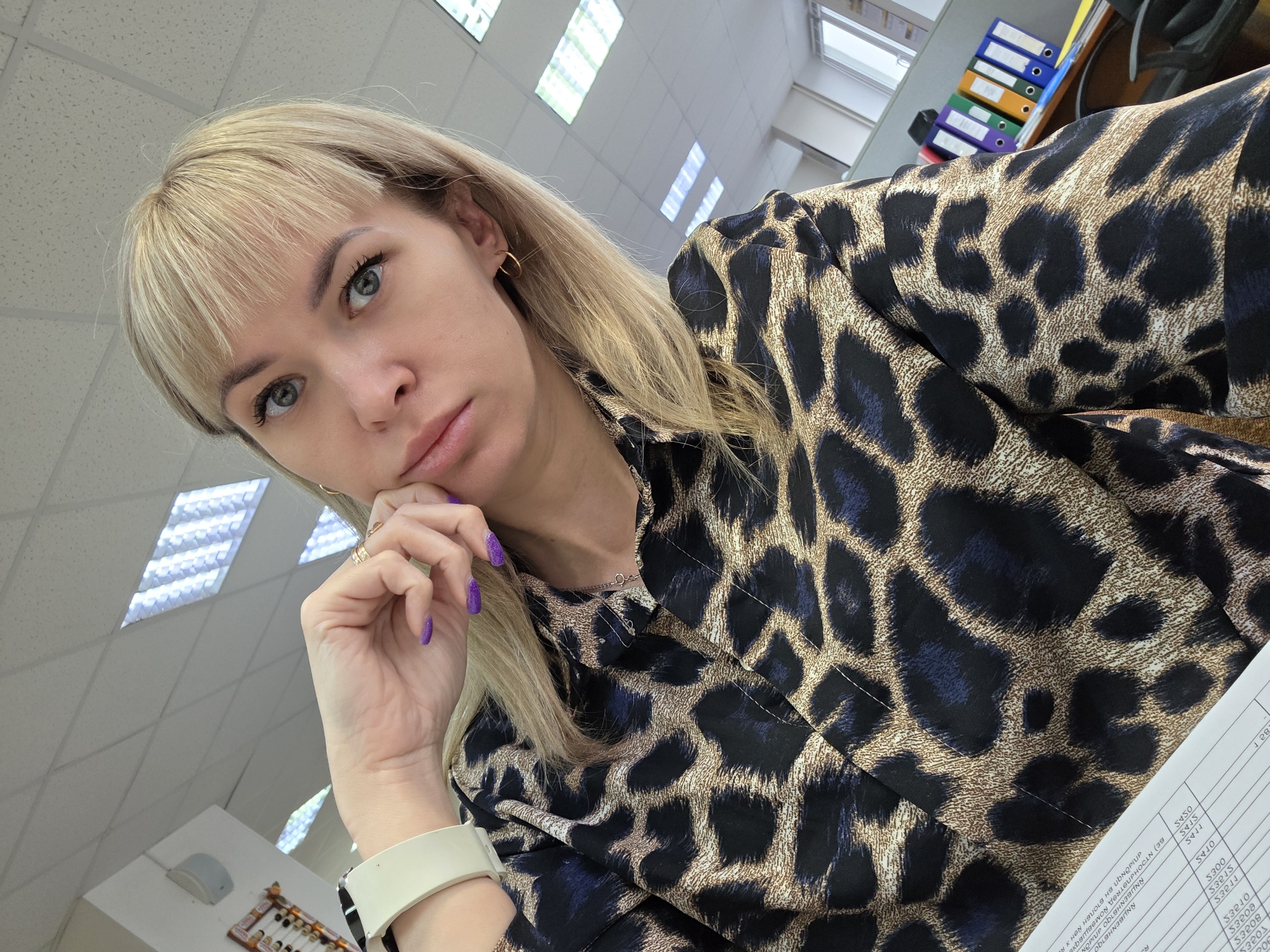 A woman with blonde hair and bangs is seated at a desk in an office. She is wearing a leopard-print shirt and has a white smartwatch on her left wrist. Her left hand is raised to her face, with her fingers touching her chin. She has purple nail polish. Her gaze is directed towards the camera.
The background includes office elements such as a ceiling with fluorescent lights and a filing cabinet with colorful binders. A piece of paper with writing on it is also visible on the desk.
The location is likely an office building in the city of Chaikovsky, Russia, indicated by the Russian text on the paper.