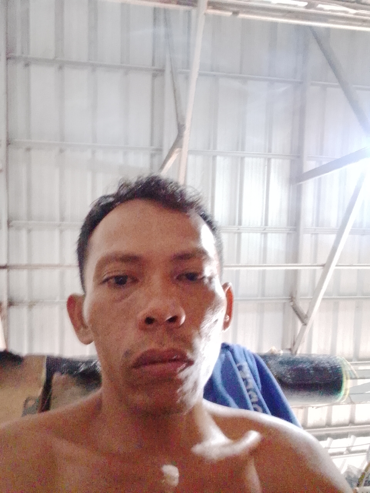 A shirtless man with short black hair is centered in a selfie-style photograph. His facial features are prominent, and he appears to be indoors. In the background, a corrugated metal structure is visible, likely a building or warehouse. A blue cloth and other unidentified objects are partially visible behind his left shoulder. The location is assumed to be Bukit Ketok, Indonesia. The lighting appears bright and somewhat diffused.