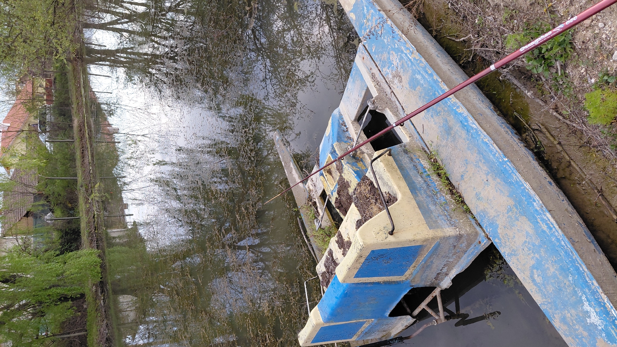 An eye-level, medium shot depicts an abandoned, decaying boat resting in a body of water, potentially a canal or narrow river. The boat is primarily blue and yellow, covered in dirt and moss, with some sections broken and decaying. A red fishing rod is resting on the boat, its tip extending towards the water. The water reflects the surrounding environment, showing trees, sky, and a building in the distance, suggesting an urban or semi-urban location. The immediate surroundings consist of overgrown vegetation and muddy banks, indicating a neglected area. There are no people visible in the scene.