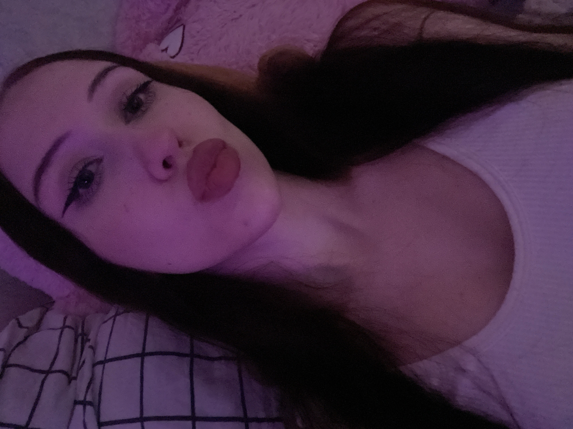 The image is a close-up, selfie-style photograph taken in what appears to be a bedroom environment, likely at night given the lighting. The subject is a young woman with dark hair, wearing a white tank top. She is looking towards the camera with her lips pursed. Her makeup includes winged eyeliner. The background is dimly lit with a pinkish-purple hue, characteristic of ambient or artificial lighting such as LED strips or colored bulbs. To the left, a portion of a black and white checkered pillow or blanket is visible. To the right, a soft, pink, plush material, possibly a stuffed animal or blanket, can be seen. The overall impression is of a casual, personal photograph. The location of Votkinsk, Russia is not indicated in the visual information provided by the image.