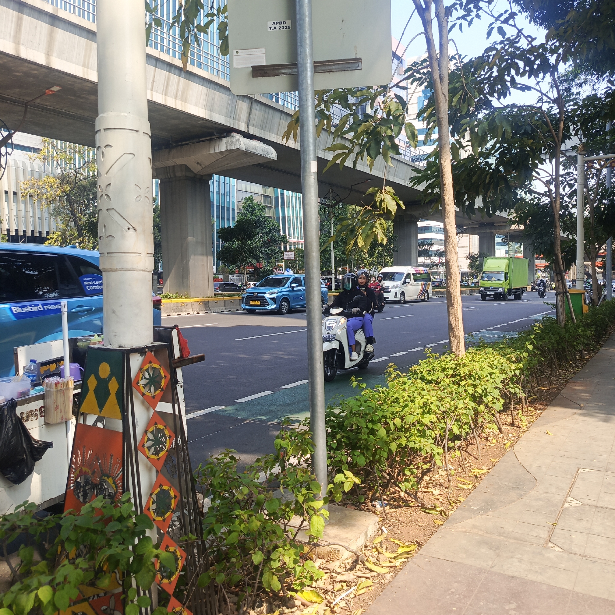 The image captures a busy urban street scene in South Jakarta, Indonesia, likely during daylight hours. In the foreground, a white pole, part of a street light or signage system, stands prominently. Attached to its base is a small street vendor's cart, adorned with colorful, geometric patterns and what appear to be small artworks. A black plastic bag rests on the left side of the cart. Behind the cart, a blue Bluebird Prime taxi is visible, parked or moving on the road.
The street itself features multiple lanes of traffic. A white scooter carrying two individuals, both wearing helmets and casual attire, travels in the right lane. The driver is looking at their phone. Further down the road, a green truck with a closed cargo container is moving forward. To the right of the road, a sidewalk with patterned tiles is present, bordered by a row of lush green bushes and trees, with fallen leaves scattered on the ground.
Overhead, a concrete elevated structure, likely an overpass or part of a transportation system, spans across the scene, supported by large concrete pillars. In the background, modern glass-fronted buildings rise, characteristic of a city environment. The sky is clear and blue. The overall impression is one of a dynamic urban environment with consistent traffic flow and commercial activity.