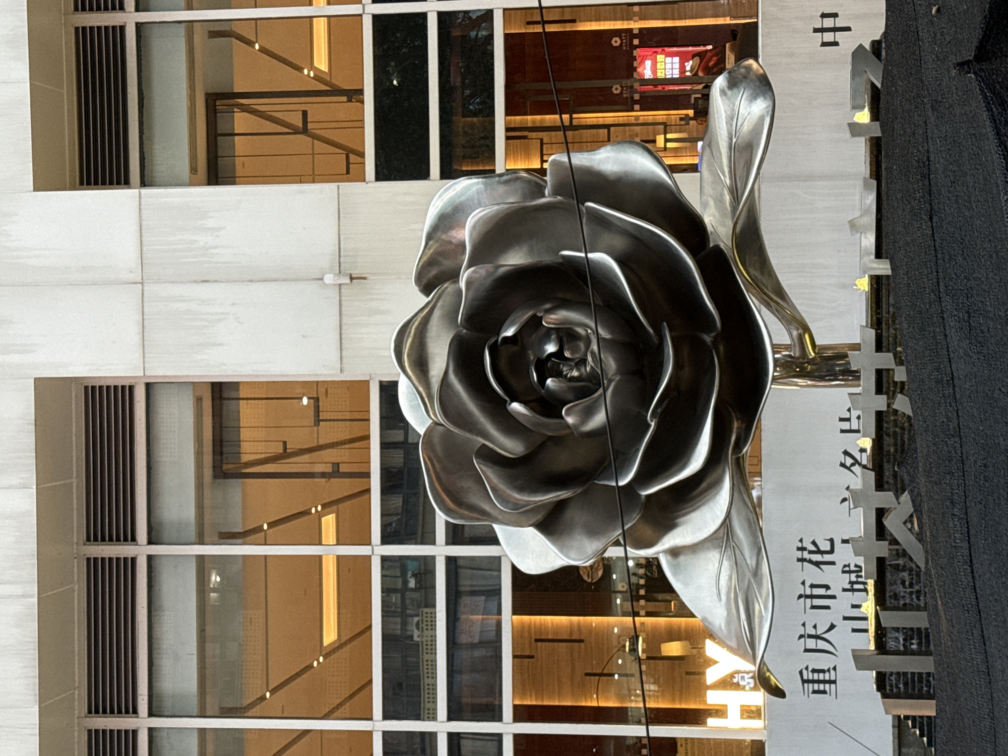 A large, metallic sculpture of a rose is prominently displayed in front of a modern building with large windows. The sculpture is silver and appears to be made of polished metal, with detailed petals and leaves. The building behind it has a facade of concrete and glass, with warm lighting visible through the windows, suggesting it is either an office building or a hotel. Below the rose sculpture, there is a white panel with Chinese characters, likely indicating the location or purpose of the sculpture. The characters appear to read "重庆市花", which translates to "Chongqing City Flower". The immediate foreground consists of a dark, textured surface, possibly a planter or a decorative base for the sculpture, with some abstract metallic elements and small yellow lights embedded within. The overall scene suggests an urban public art installation in Yuzhong District, China.