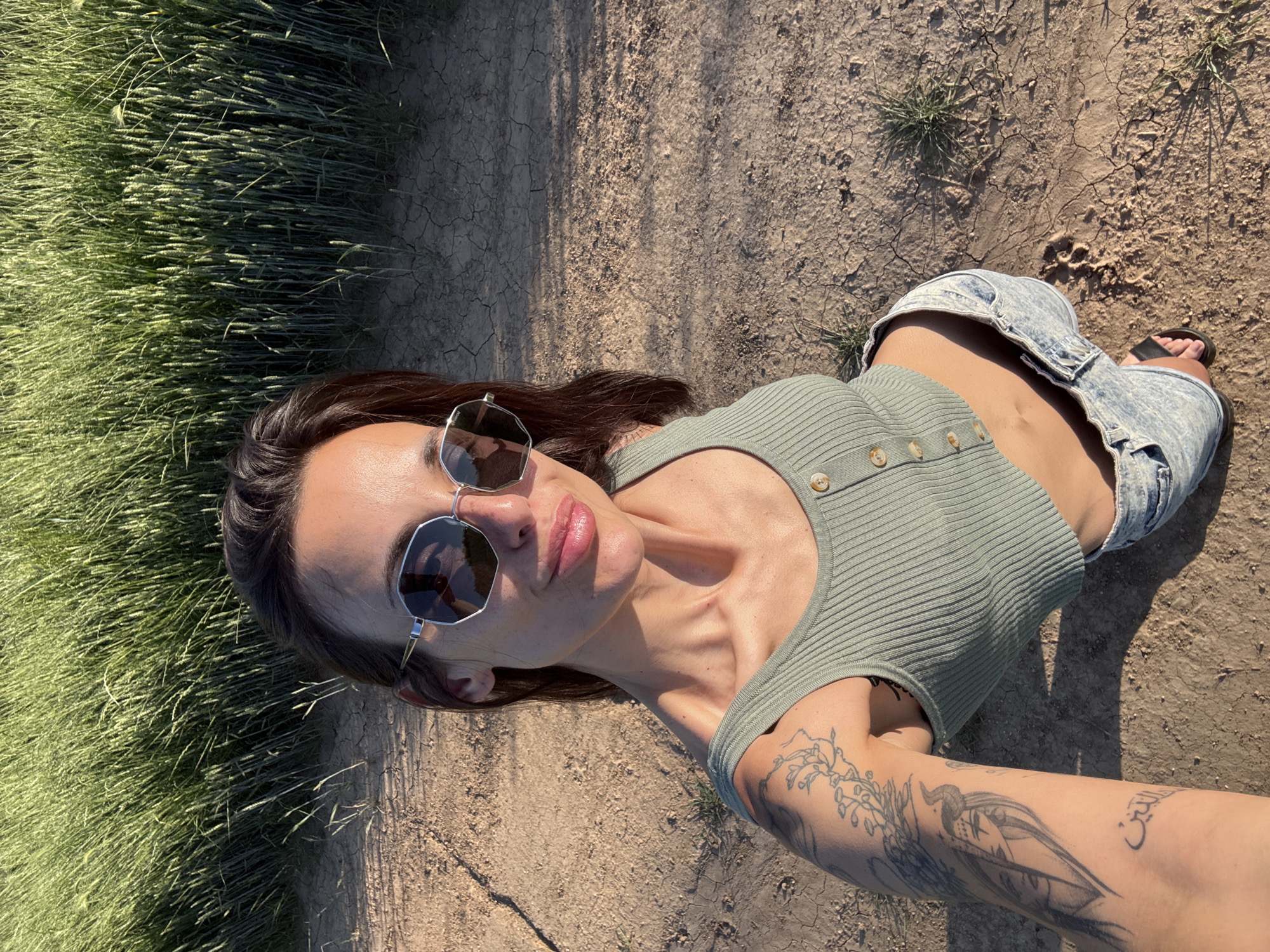 A medium-shot photograph from an elevated viewpoint depicts a woman positioned on cracked, dry soil, bordering a grassy area to her left. The woman is wearing sunglasses, a green ribbed top with buttons, and denim shorts. A black sandal is visible on her right foot. Her right arm, featuring multiple tattoos, is extended towards the camera. The location is not identifiable as Pleven, Bulgaria, based on the image.