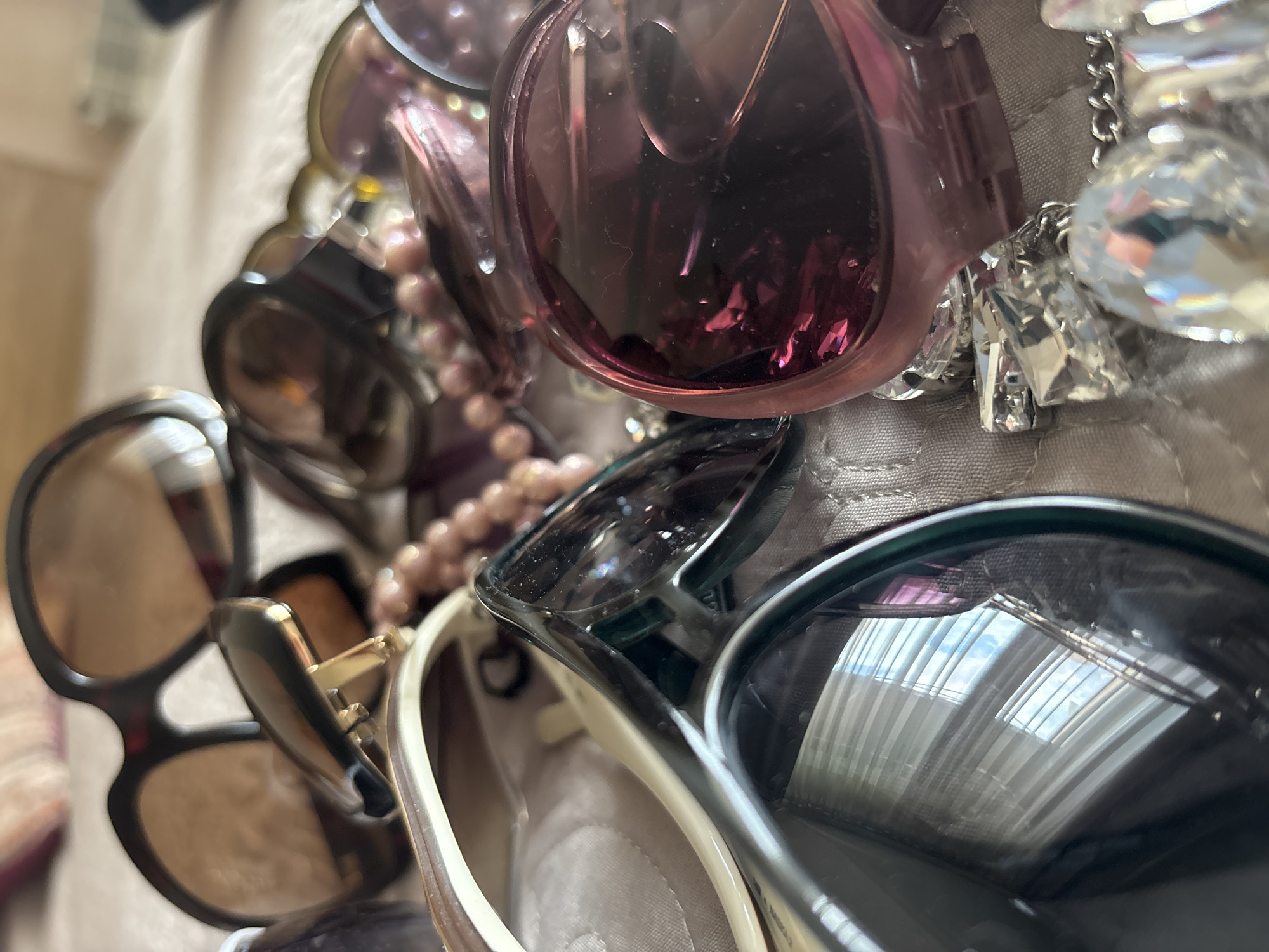 The image shows a close-up of a pile of sunglasses and costume jewelry on a textured, possibly quilted, fabric surface. There are several pairs of sunglasses with varying frame colors and shapes, including brown, black, and pink. The jewelry consists of strings of beads and items with clear, possibly crystal, elements. The reflection in one of the sunglass lenses shows a window with white blinds, revealing that the photo was taken indoors. There is no information in the image that relates to the city of Minsk, Belarus.