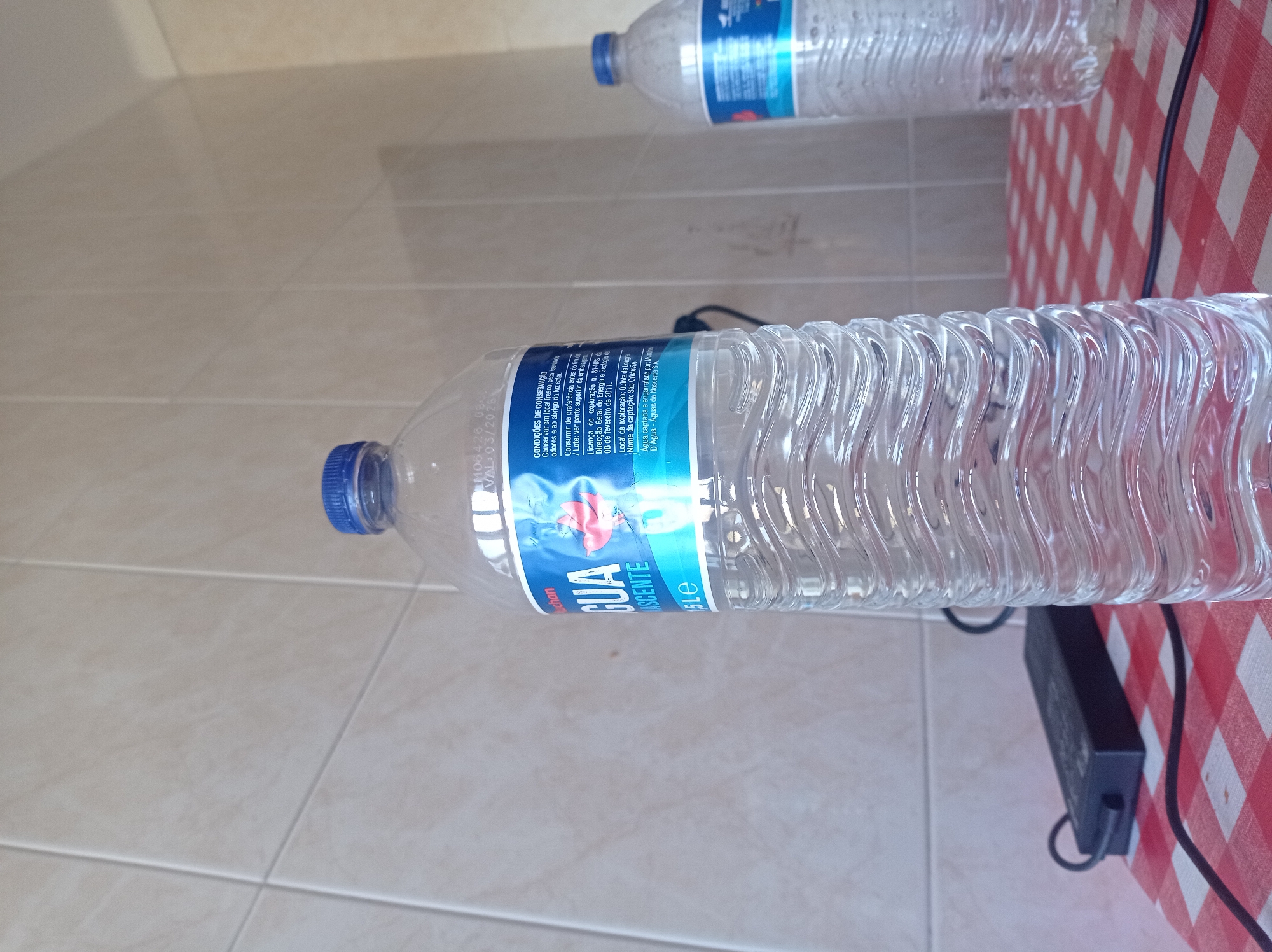 The scene depicts an interior setting. A large, clear plastic water bottle with a blue screw-top cap and a blue "Auchan AGUA NASCENTE 1,5Le" label is prominently featured in the foreground, partially filled with clear liquid. The label displays text in Portuguese, including a visible best-before date of March 2025. To the left of this bottle, on a red and white checkered tablecloth, lies a black rectangular electronic power adapter with a connected black cable. In the mid-ground, a second, partially obscured clear plastic water bottle with a blue cap and label stands, exhibiting condensation. The background consists of a light beige tiled wall. No individuals, actions, or interactions are visible. Location: City of Unknown, Unknown.
