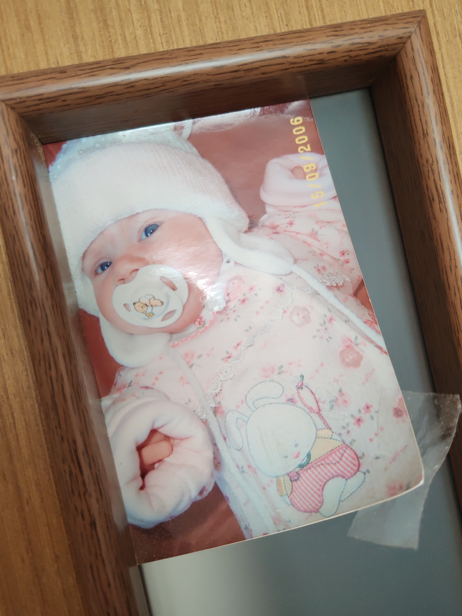 The media file presents a slightly tilted view of a framed photograph, showcasing an infant. The photograph is set within a brown wooden frame featuring a prominent wood-grain texture. A gray matte borders the printed image.
The central subject of the photo is a fair-skinned baby with clear blue eyes, depicted in a serene, possibly posed, state. The baby wears a cozy white knitted hat that covers its head and ears. A white pacifier, decorated with a small teddy bear design, is in the baby's mouth. The infant is dressed in a light pink sleeper suit or overall, adorned with a delicate floral pattern of small flowers and leaves, and accented with white lace trim. On the lower right side of the suit, a white bunny appliqué is visible, dressed in pink striped overalls. The baby's small, bundled hands are partially seen.
A date stamp, "15/09/2006", is visible in yellow text, oriented vertically, on the upper right side of the photograph. A small piece of clear tape or plastic is affixed to the bottom right corner of the photograph, extending slightly onto the gray matte. The framed picture appears to be resting on or against a surface with a light wooden texture, similar to the frame, indicating an indoor, domestic setting. While the image itself provides no direct geographical cues, the provided context places this scene potentially in Saint Petersburg, Russia. The overall mood conveyed is one of peaceful innocence.