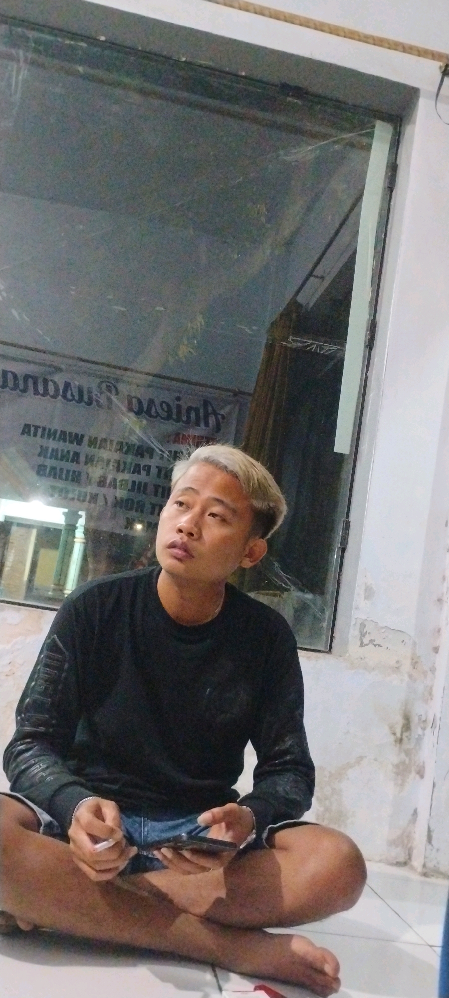 In Mumbulsari, Indonesia, a young man with bleached blonde hair sits cross-legged on the floor in front of a large, dirty window. He is wearing a black long-sleeved shirt and denim shorts. He holds a smartphone in his right hand and a cigarette in his left hand. The window reflects a dimly lit interior with signs that appear to be in Indonesian. The wall to the right of the window is peeling and discolored, suggesting an older or neglected building. The overall lighting is dim, with the man's face illuminated from the front, casting shadows.