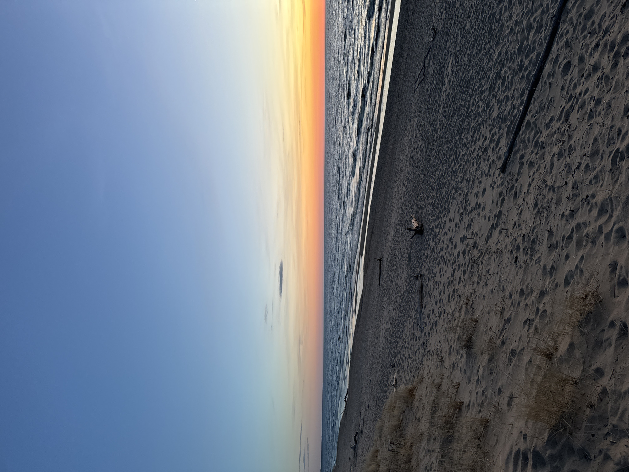 The image depicts a beach scene in Garciems, Latvia. The sky transitions from blue to orange near the horizon, indicating sunrise or sunset. The sea is visible with small waves. The sandy beach in the foreground is dark and damp, showing footprints and some sparse vegetation. A piece of driftwood lies on the sand. No people are visible in the scene.