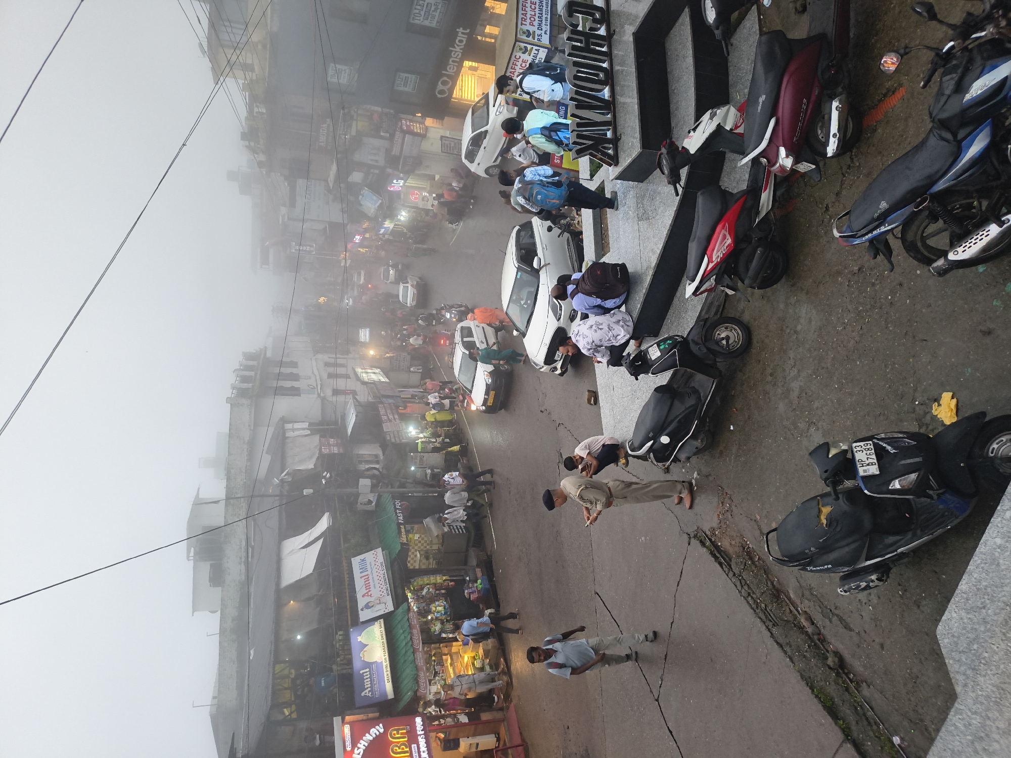 The image depicts a misty street scene in Dharamshala, India, likely a commercial area with shops and vehicles. In the foreground, several scooters and motorcycles are parked. One scooter, a black and blue model, has a license plate visible that reads "HP 33 B 6893". A person in a light blue shirt and dark pants is standing near a scooter on the right side of the image. Further up the street, more people and vehicles are visible, including cars and scooters, navigating through the dense fog. Several shops with illuminated signage are present on both sides of the street. Notably, there are signs for "Amul Milk" and "Amul", as well as what appears to be a fast-food establishment. People are seen walking and gathered in small groups. A police officer in uniform is standing and interacting with someone on the right side of the image. The overall atmosphere is one of a bustling street scene partially obscured by mist.