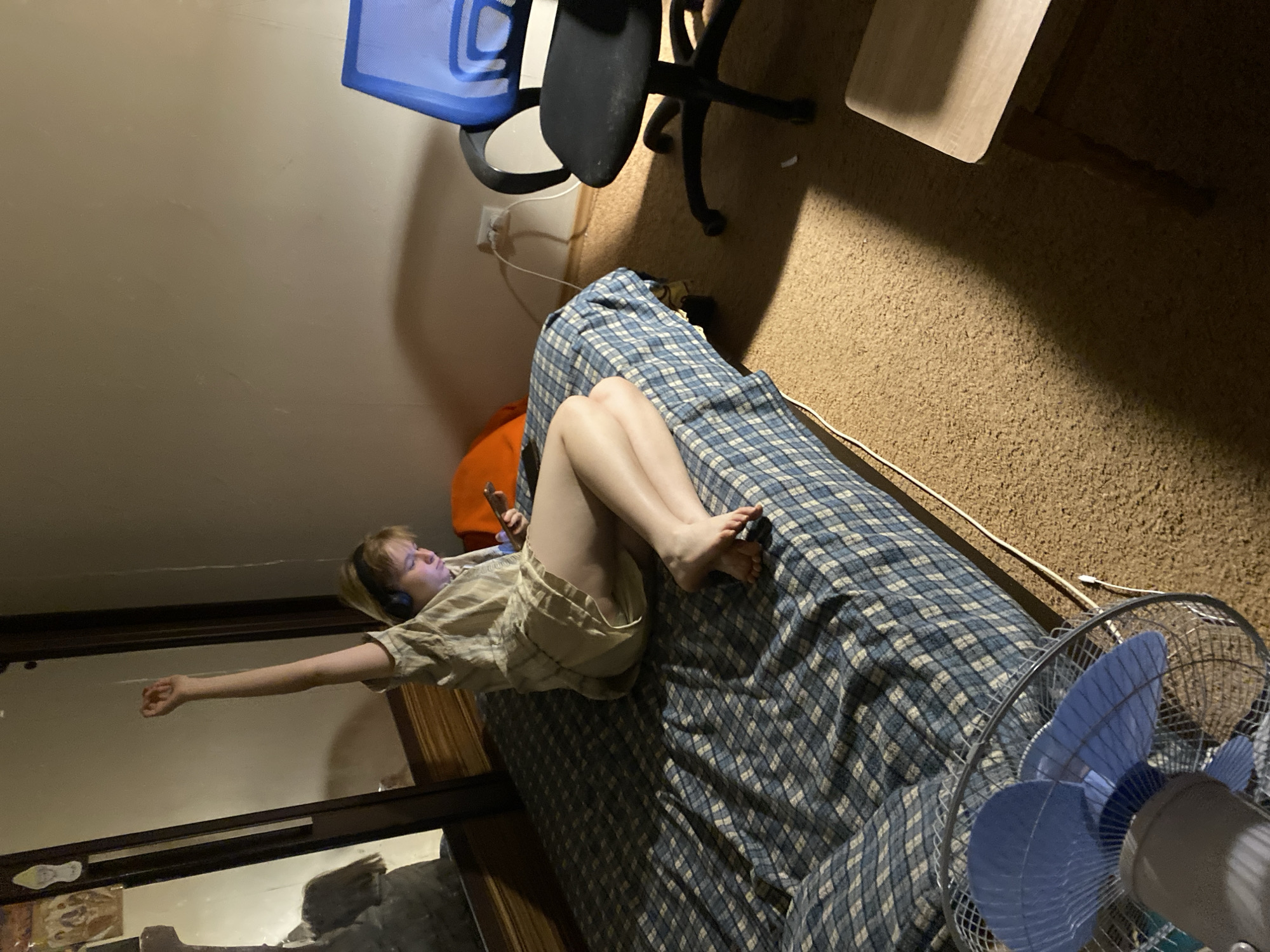 In a dimly lit room, a person with blonde hair, wearing headphones and a light-colored shirt, sits on a bed with a blue and white checkered comforter. The person is holding a mobile phone and has their right arm raised in the air. Their legs are crossed, with their right leg resting on top of their left. Next to the bed, an orange pillow is visible. To the right of the bed, a blue mesh office chair with a black base is positioned on a carpeted floor. A white electrical cord runs from a wall outlet to an unseen device. In the foreground, a blue oscillating fan with a metal grill sits on the carpet. A portion of a wooden stool is visible in the lower right corner. The room appears to be a bedroom, possibly in a residential area of Chornomorsk, Ukraine.