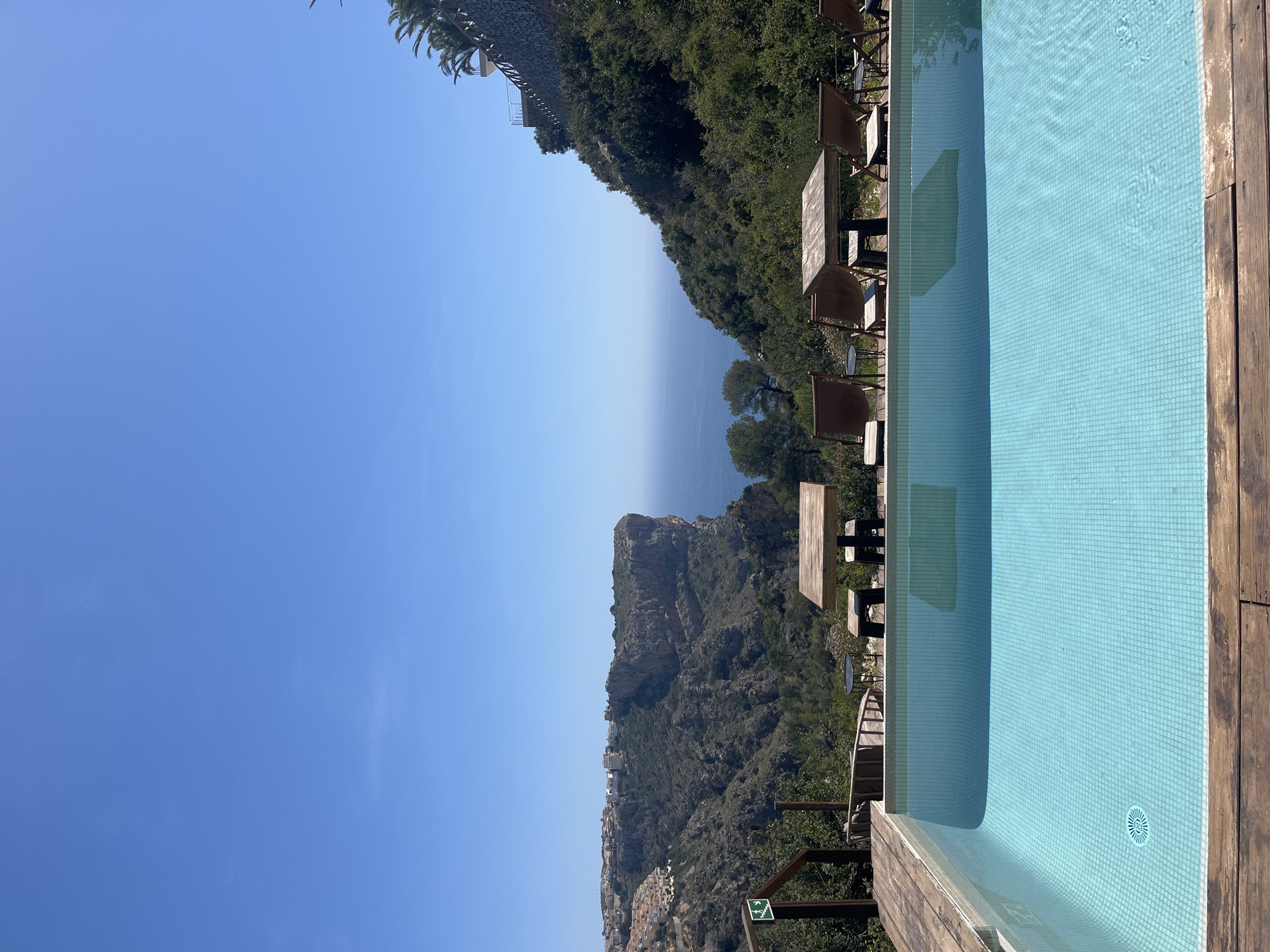 A landscape shot of Urbanització El Cim Del Sol, Spain, presents a blue sky dominating the left, meeting a body of water – likely the Mediterranean Sea – with a gradient horizon. A lush, green cliffside descends towards the sea, featuring tables and chairs arranged along the edge. To the right, a light blue tiled swimming pool with wooden decking occupies the foreground, reflecting the surrounding environment. The overall scene suggests a high vantage point with a coastal view.