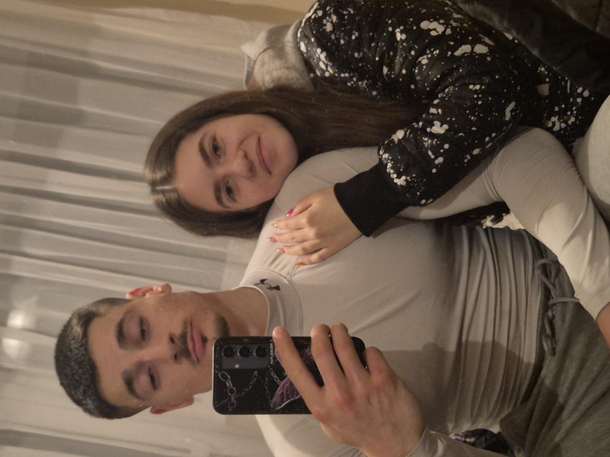 A man and a woman are indoors, likely in a home. The man is holding a smartphone with a black case, taking a selfie. The woman is lying on or behind the man, resting her head on his shoulder. She is wearing a black and white patterned hoodie. The man has short dark hair and is wearing a light gray shirt. A curtain is visible in the background. There is no indication of the location being in Pavlikeni, Bulgaria.